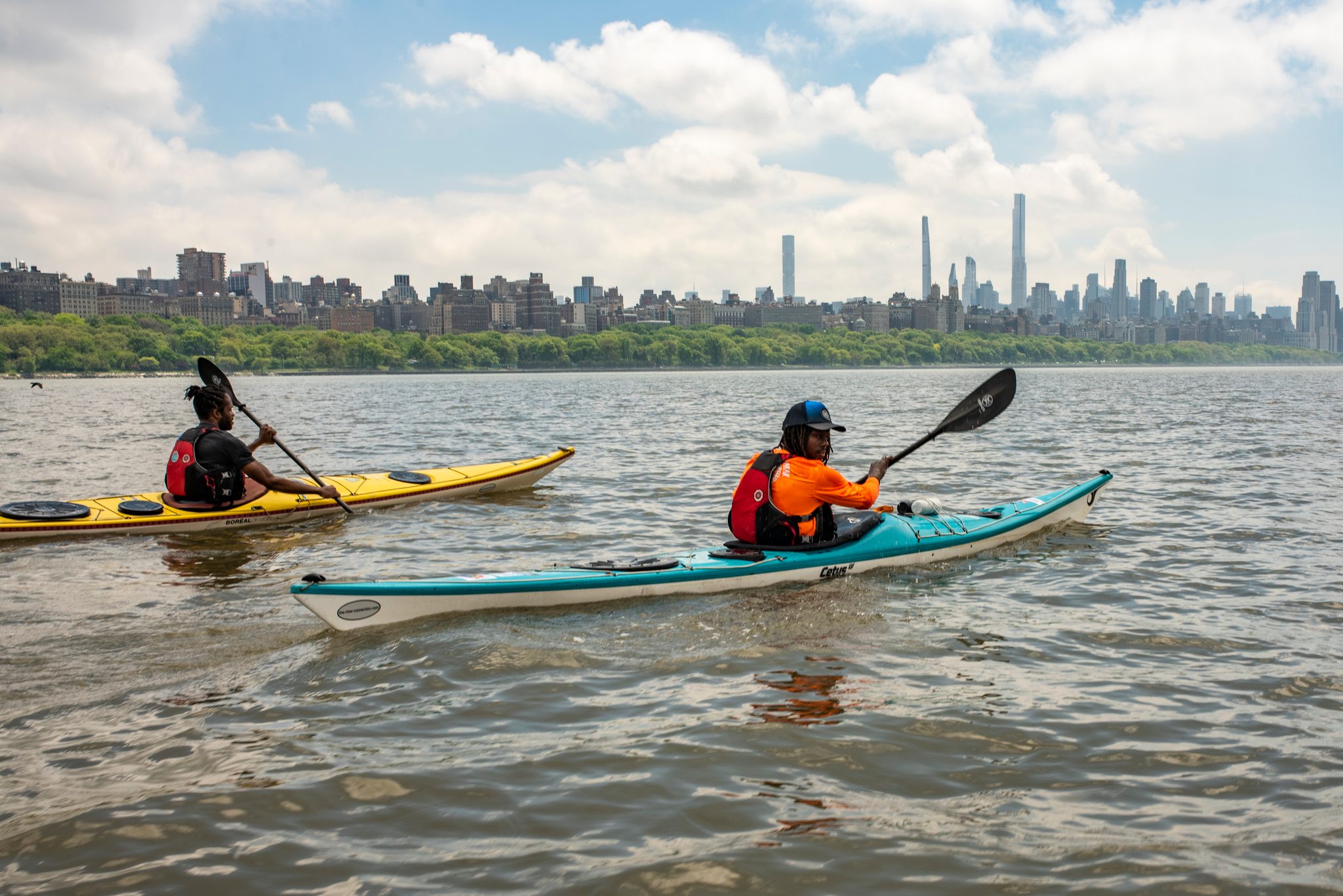 Closer now: paddles up, the orange jacket in front, the whole city stacked along the far bank.