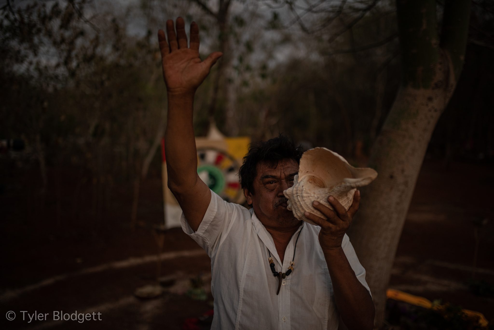 A man raises one hand to the sky and sounds the conch shell.