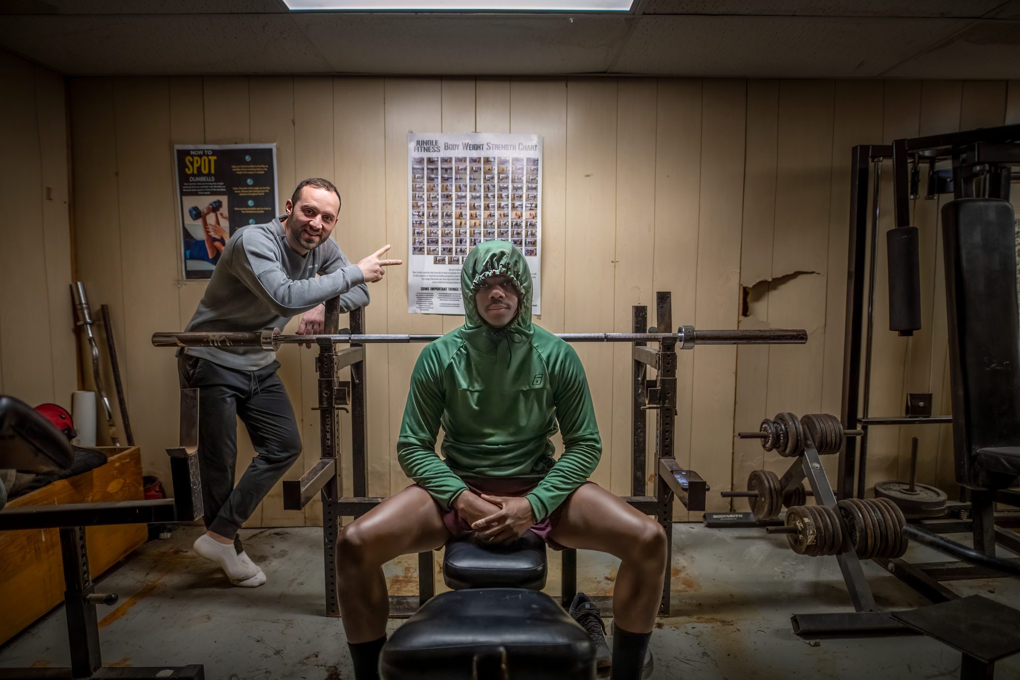 A cramped basement gym, barbells and a weight bench, two men posing mid session in the dojo.