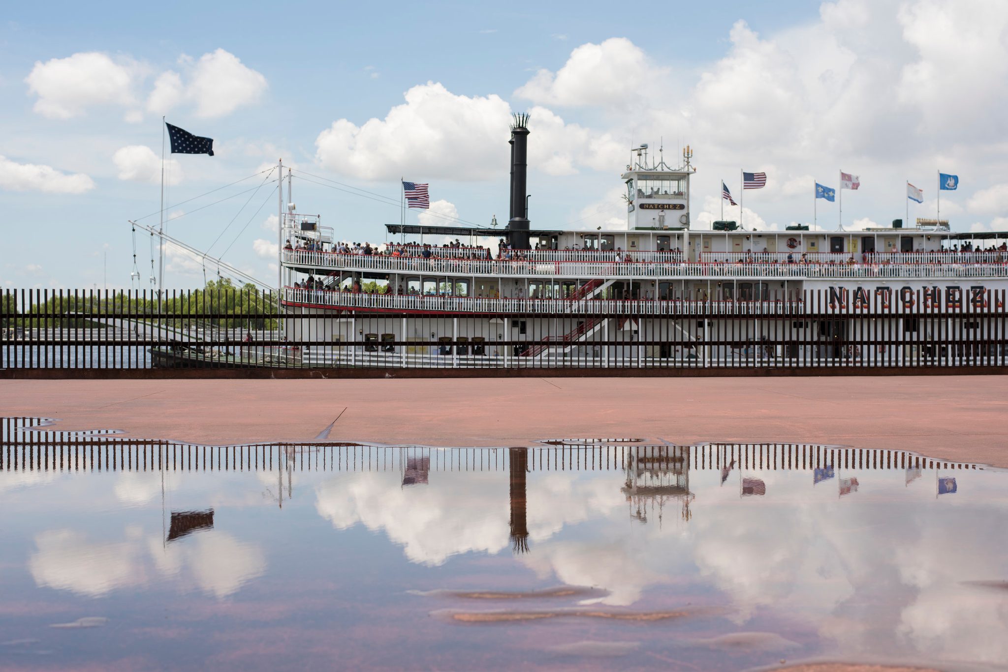 The Natchez waits at the dock, flags flying, the whole boat mirrored in a puddle.