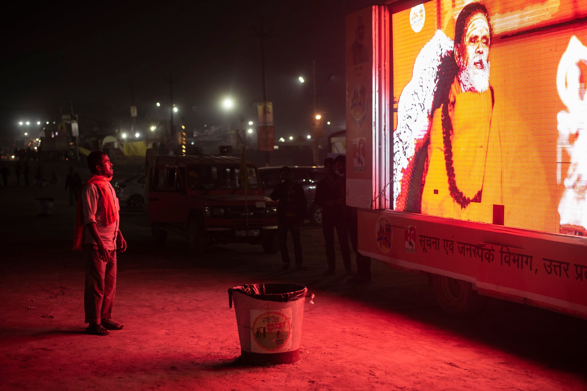 A man stands, captured in crimson light, downloading the teachings from the glowing roadside sadhu. Here at Kumbh consciousness moves. It is pulled and pushed. For some it shrinks and others it expands.