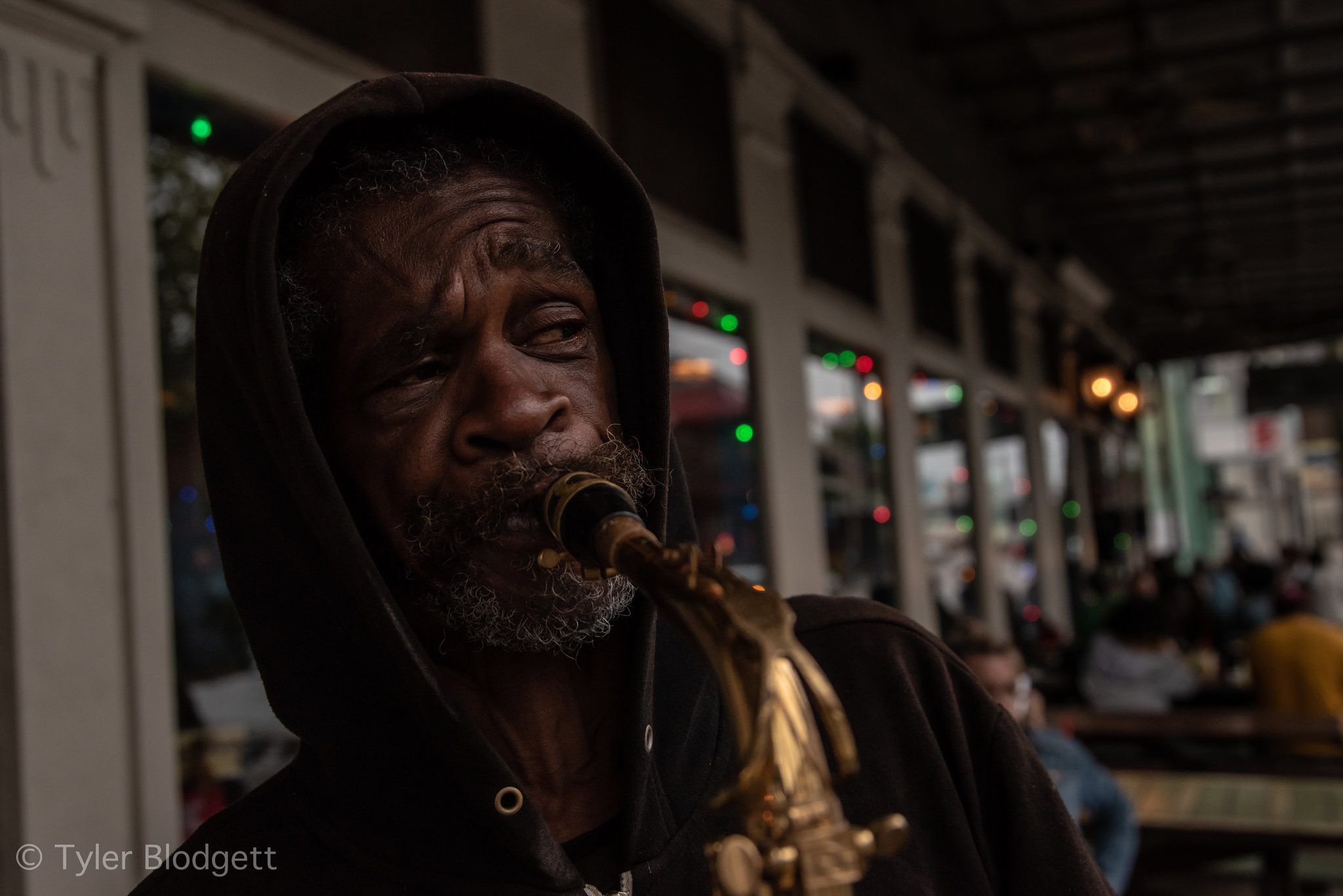 An old man in a hood plays saxophone under the gallery, eyes half closed.
