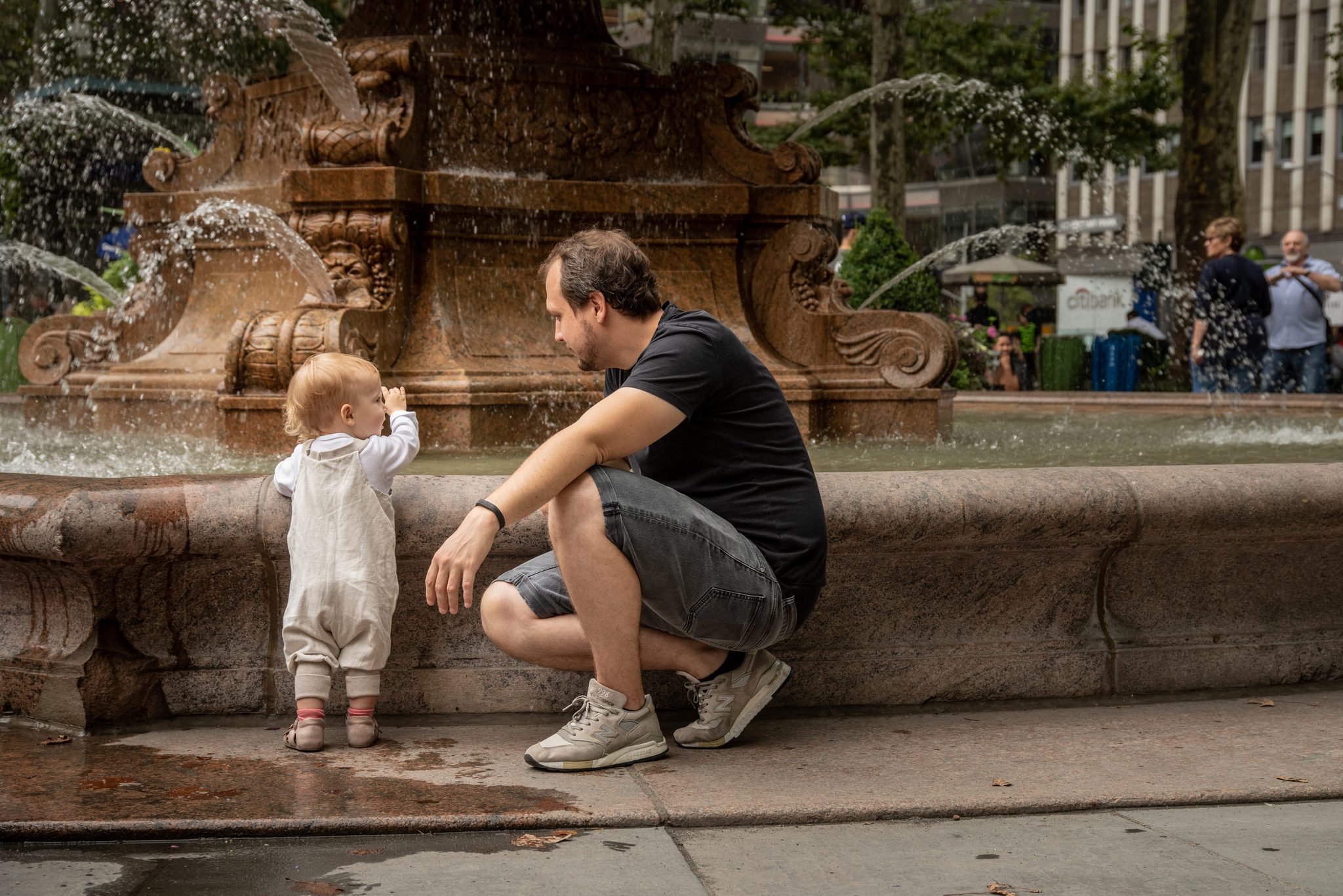A father crouches at the fountain's edge as his toddler reaches for the spray.