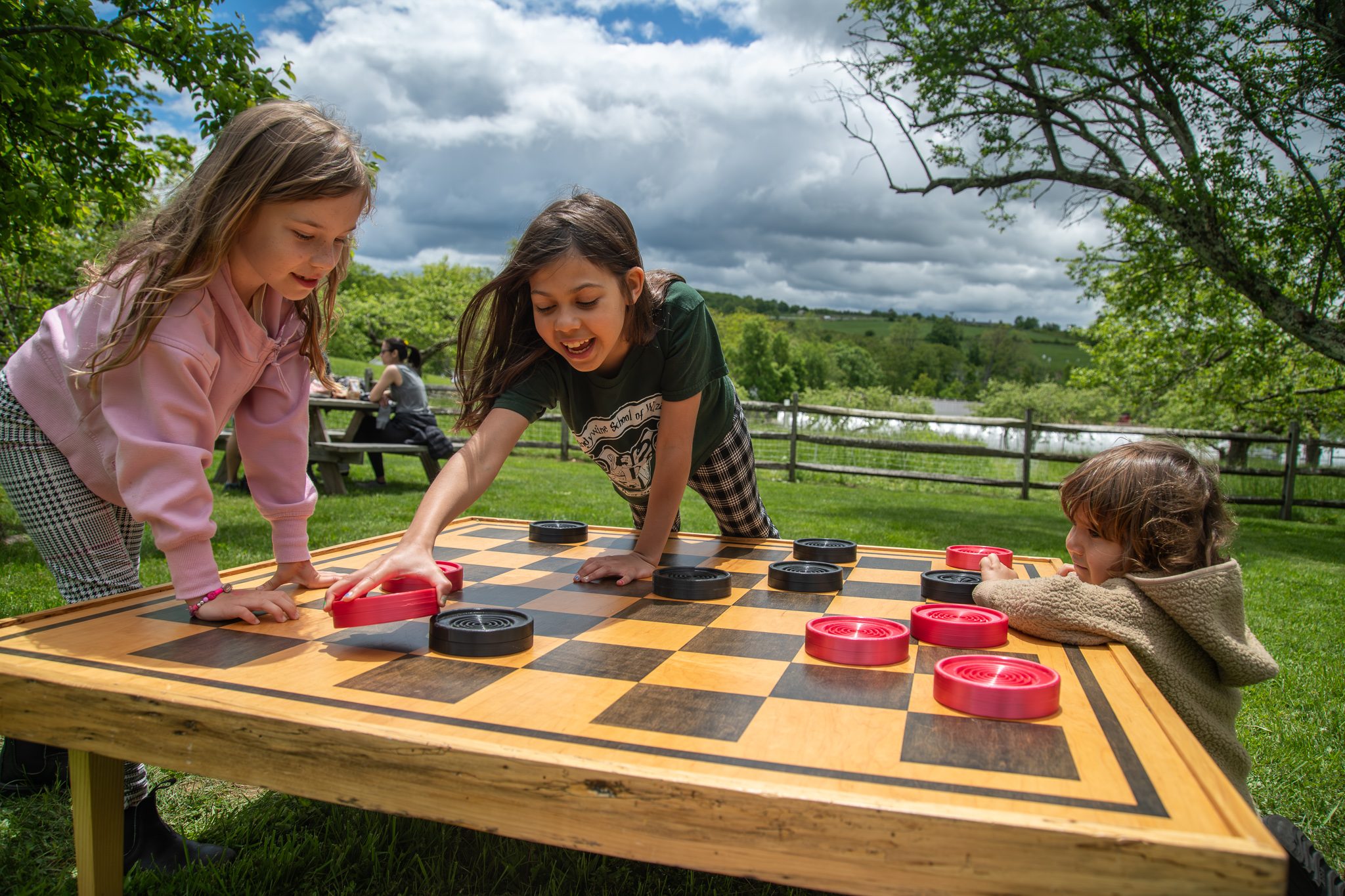Two girls reach across a giant checkerboard while a small one watches from the edge.