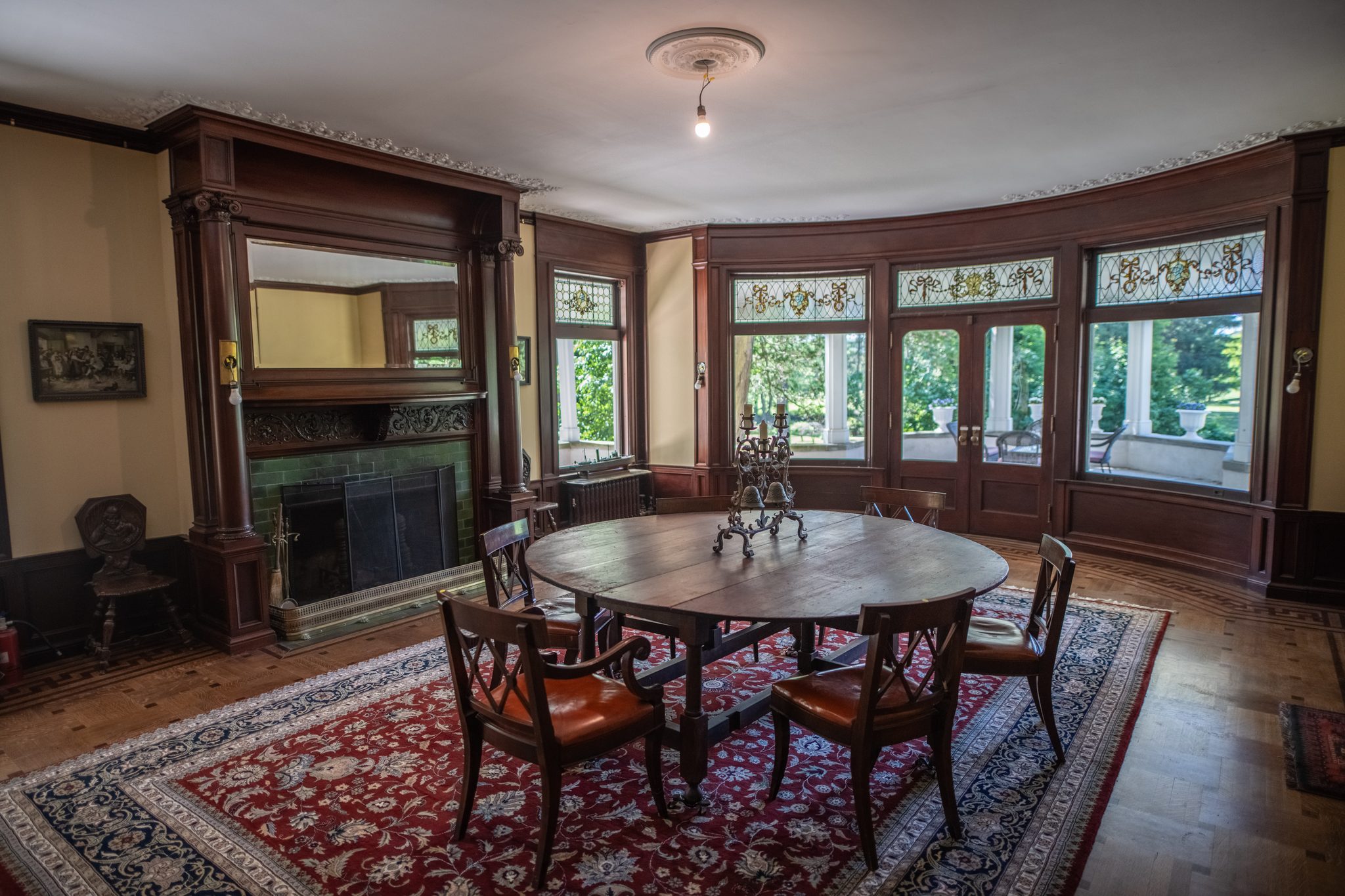A dining room with a carved mantel and green tile fireplace, the curved bay of windows opening onto the porch.