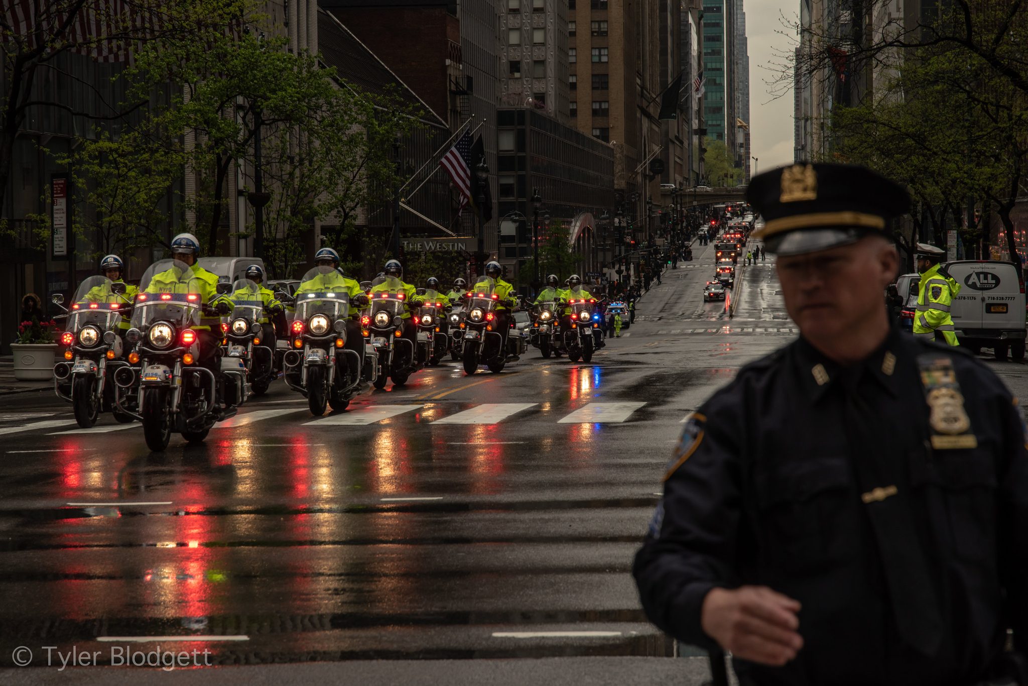 A motorcycle escort rolls up the wet avenue as an officer steps into the frame.