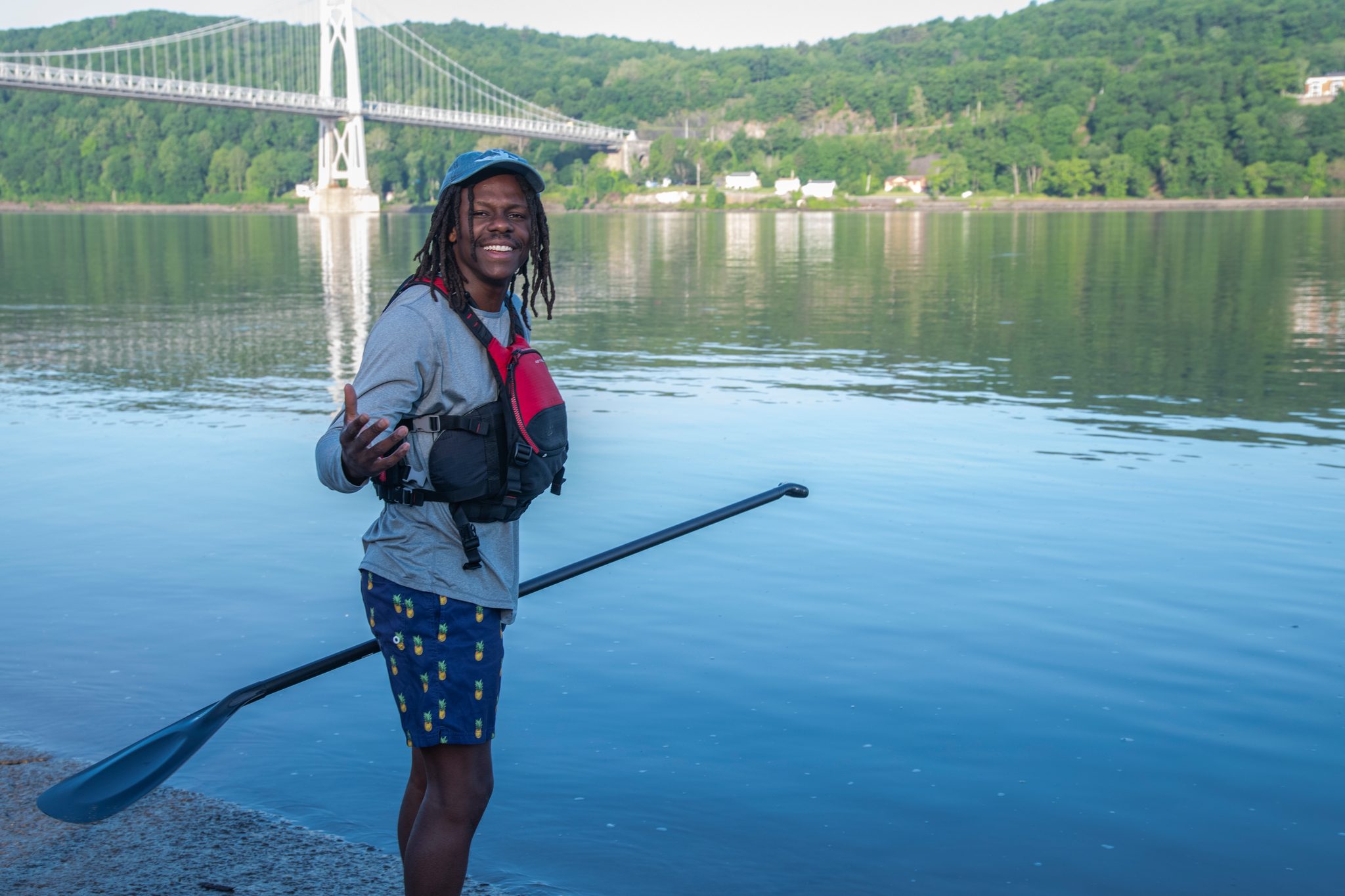 Chev stands at the water with paddle in hand, a suspension bridge and green hills behind him.
