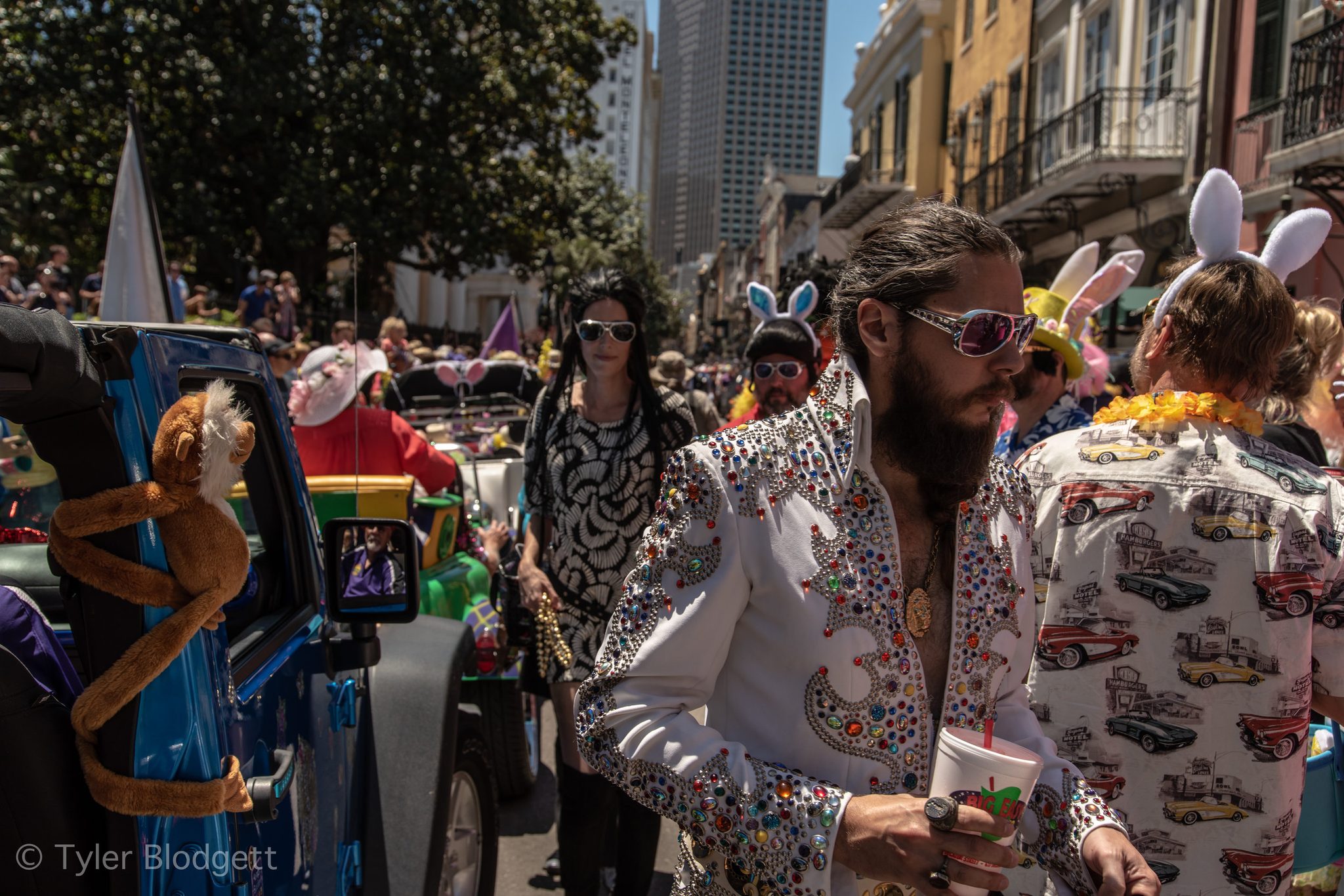 A bearded man in a rhinestone jumpsuit walks beside a monkey riding the car.