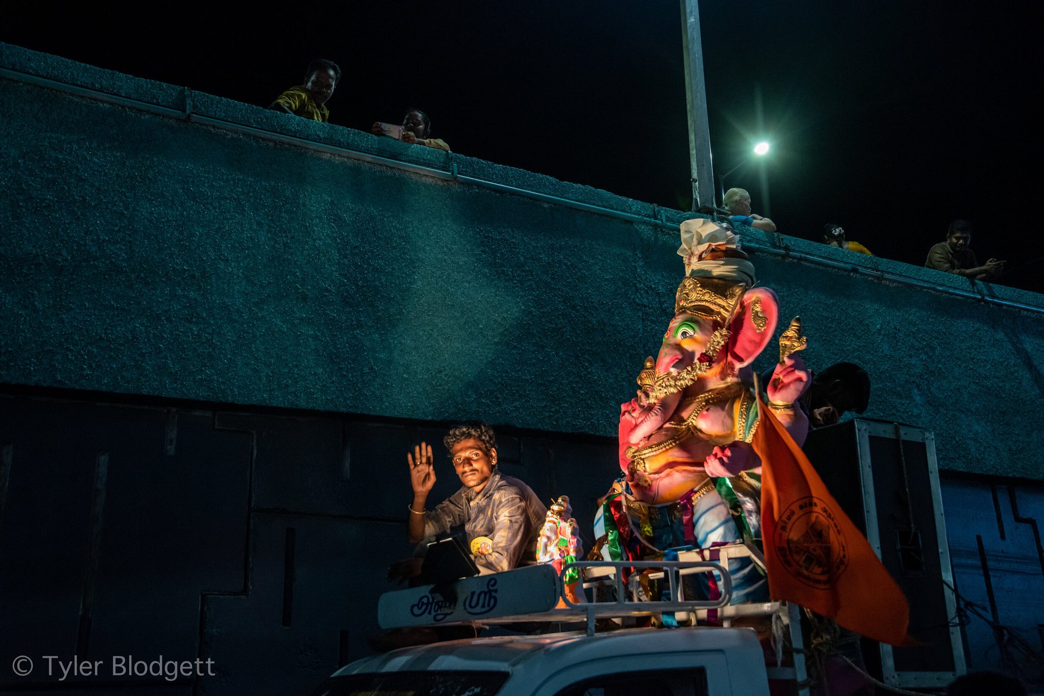 A young man rides beside Ganesha through the night, the idol glowing pink and gold above the truck.