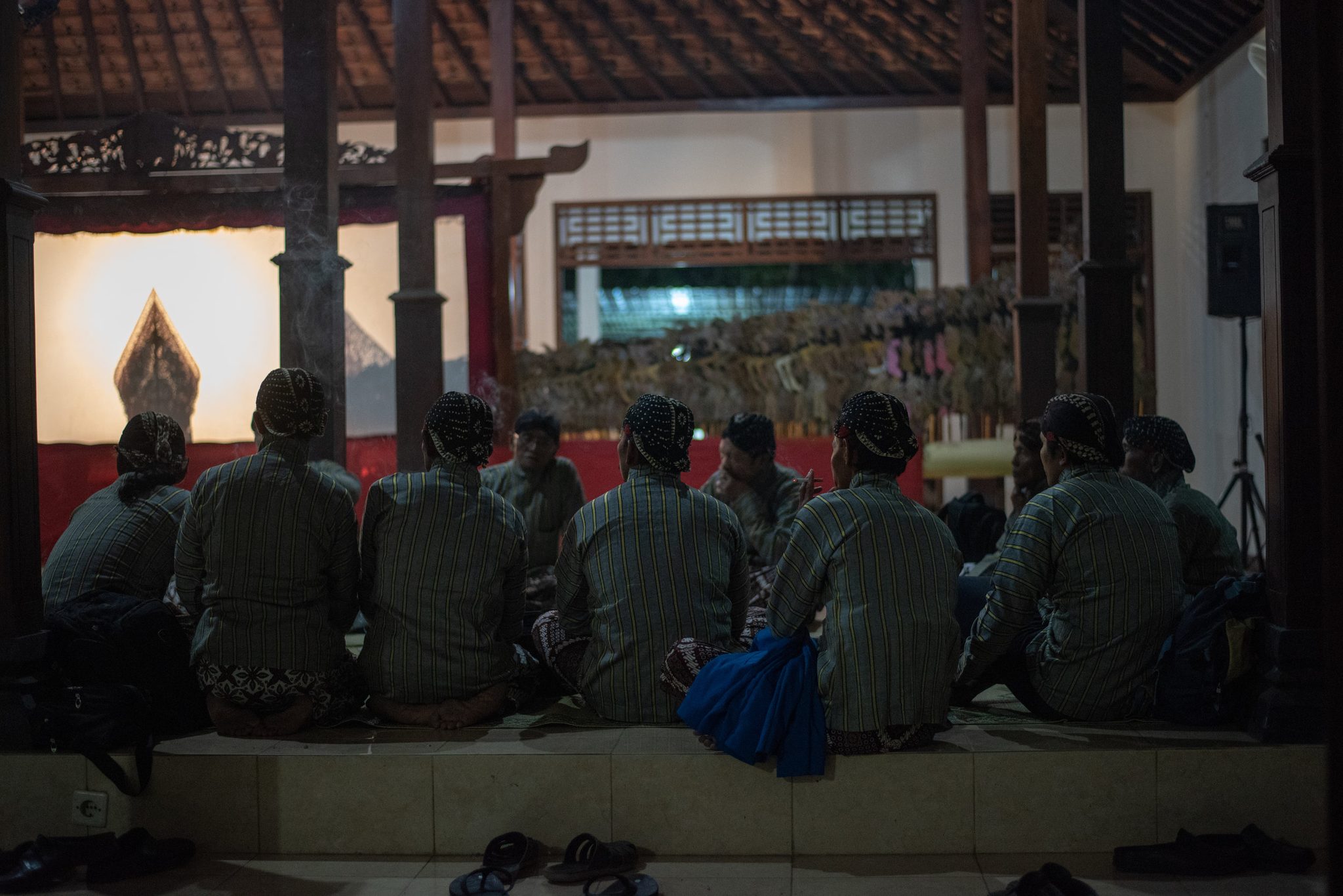 The musicians sit shoulder to shoulder facing the screen, their sandals left at the edge of the floor.