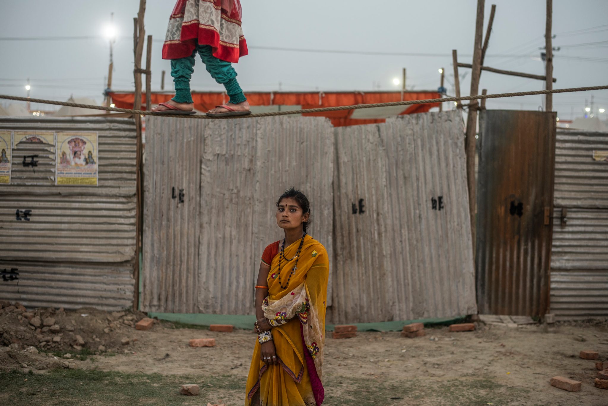 A young girl balances on a tight rope while another stands stoically below to collect money from bystanders.