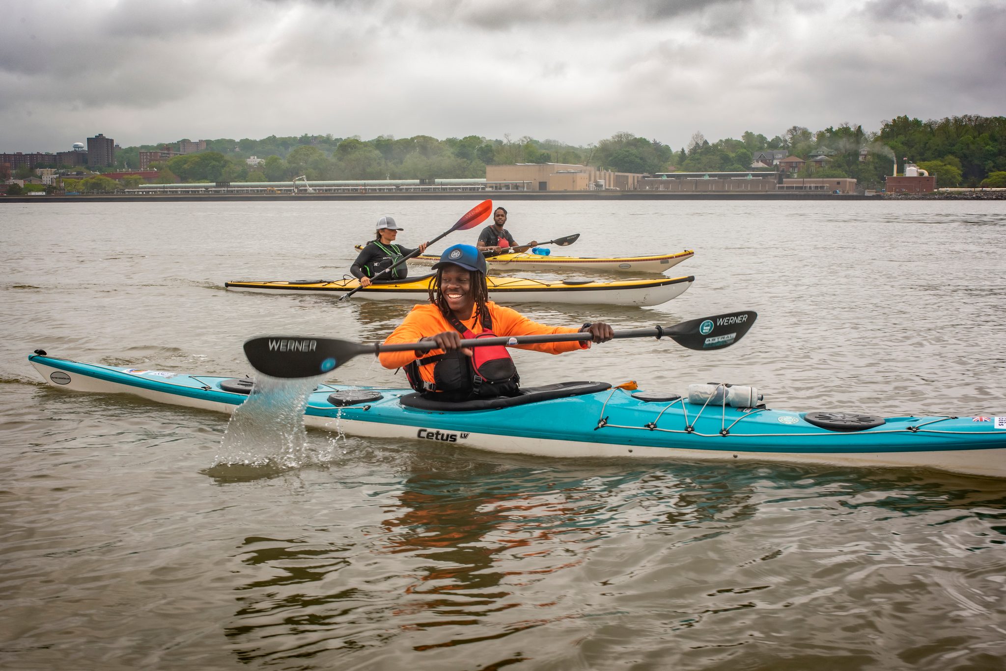 A paddler in an orange jacket leads the pack, the rest of the group strung out in yellow and red behind.