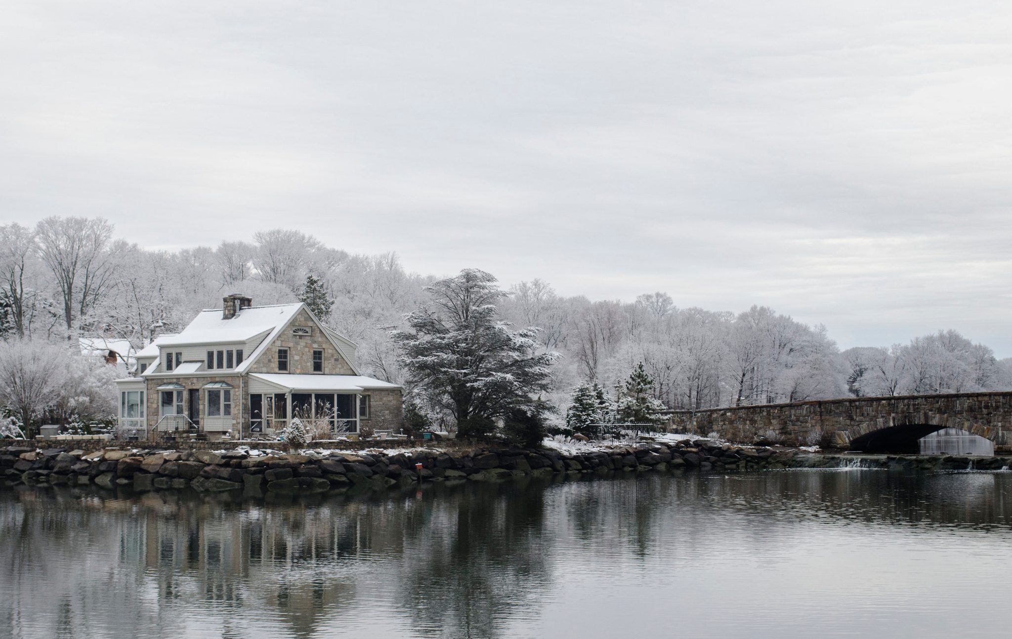 Snow blankets the stone house and the bridge beside the still water.