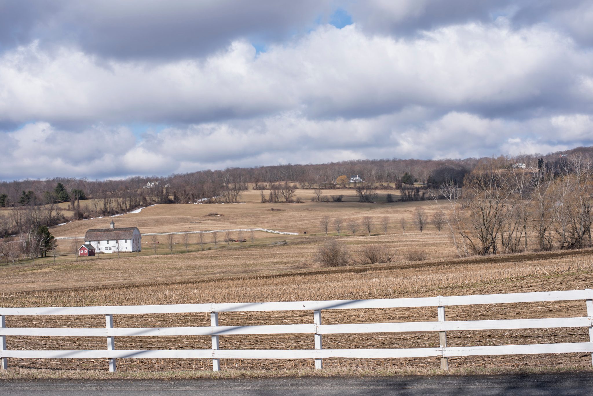 A white farmhouse sits alone across the winter fields, a white rail fence in front.