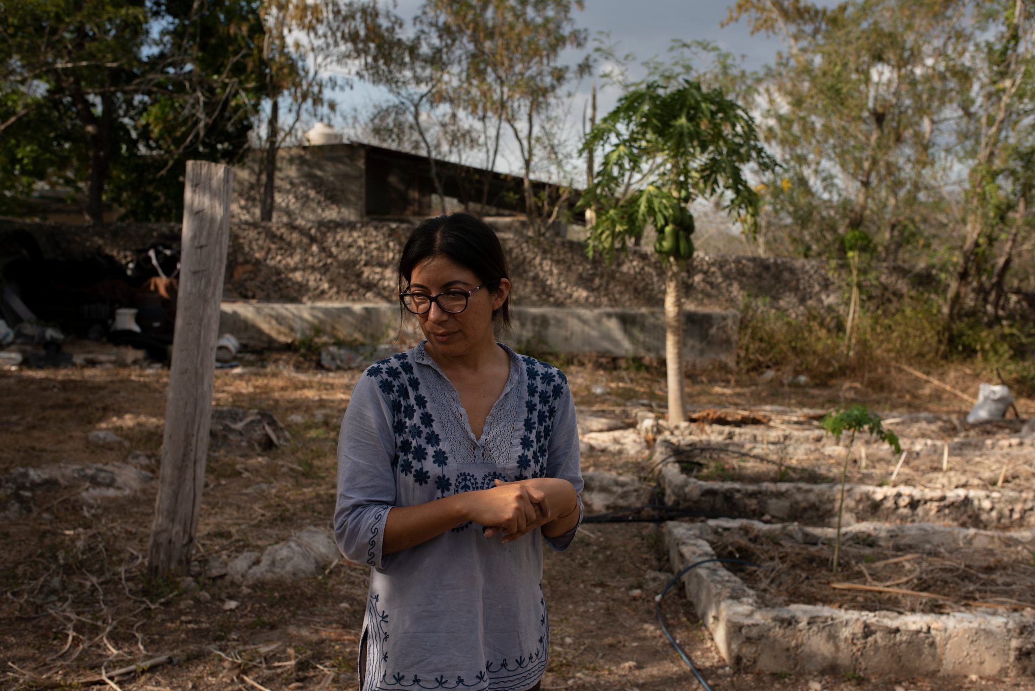 A woman in an embroidered blouse stands among the raised garden beds.