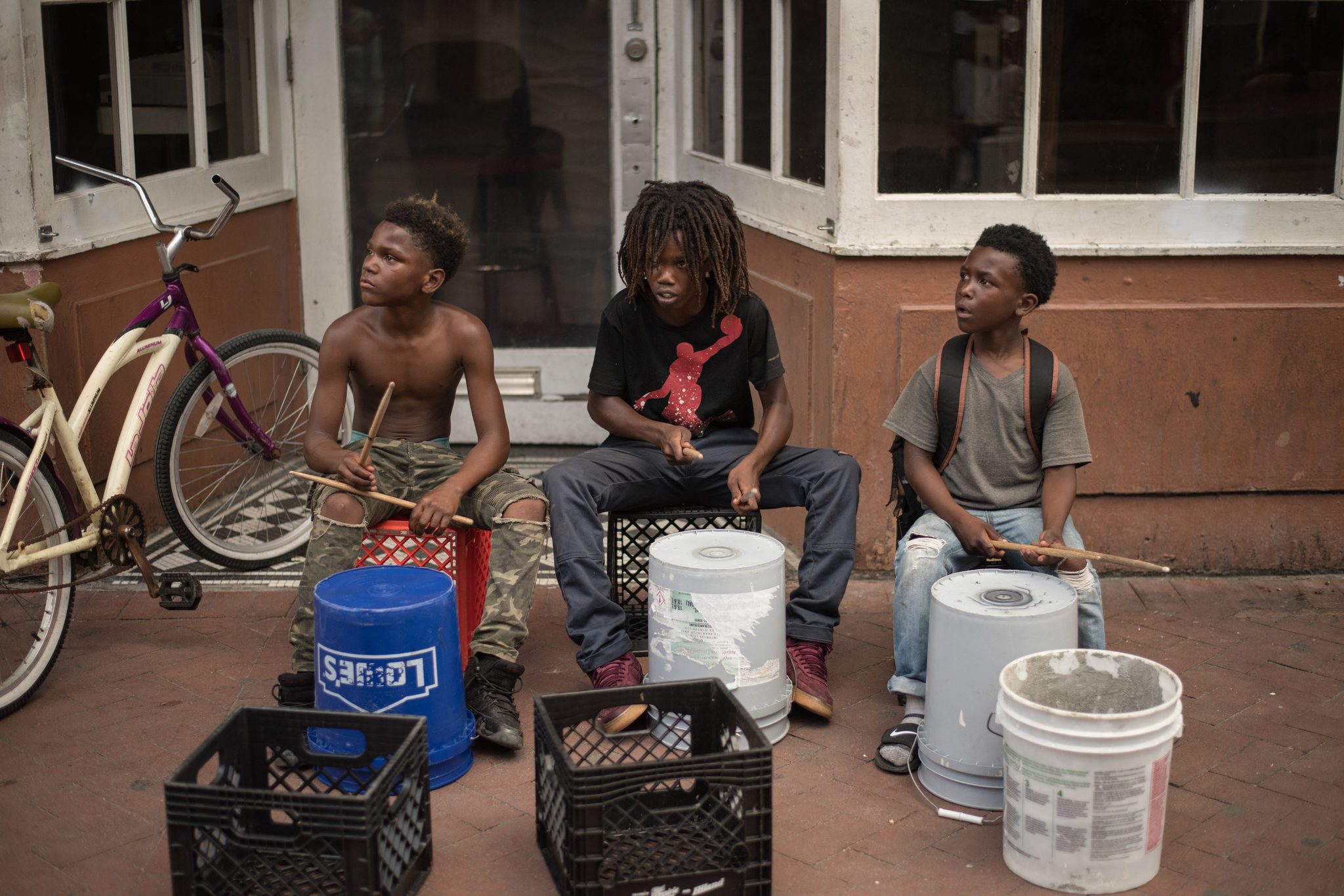 Three boys drum on buckets and crates on the sidewalk, sticks in motion.
