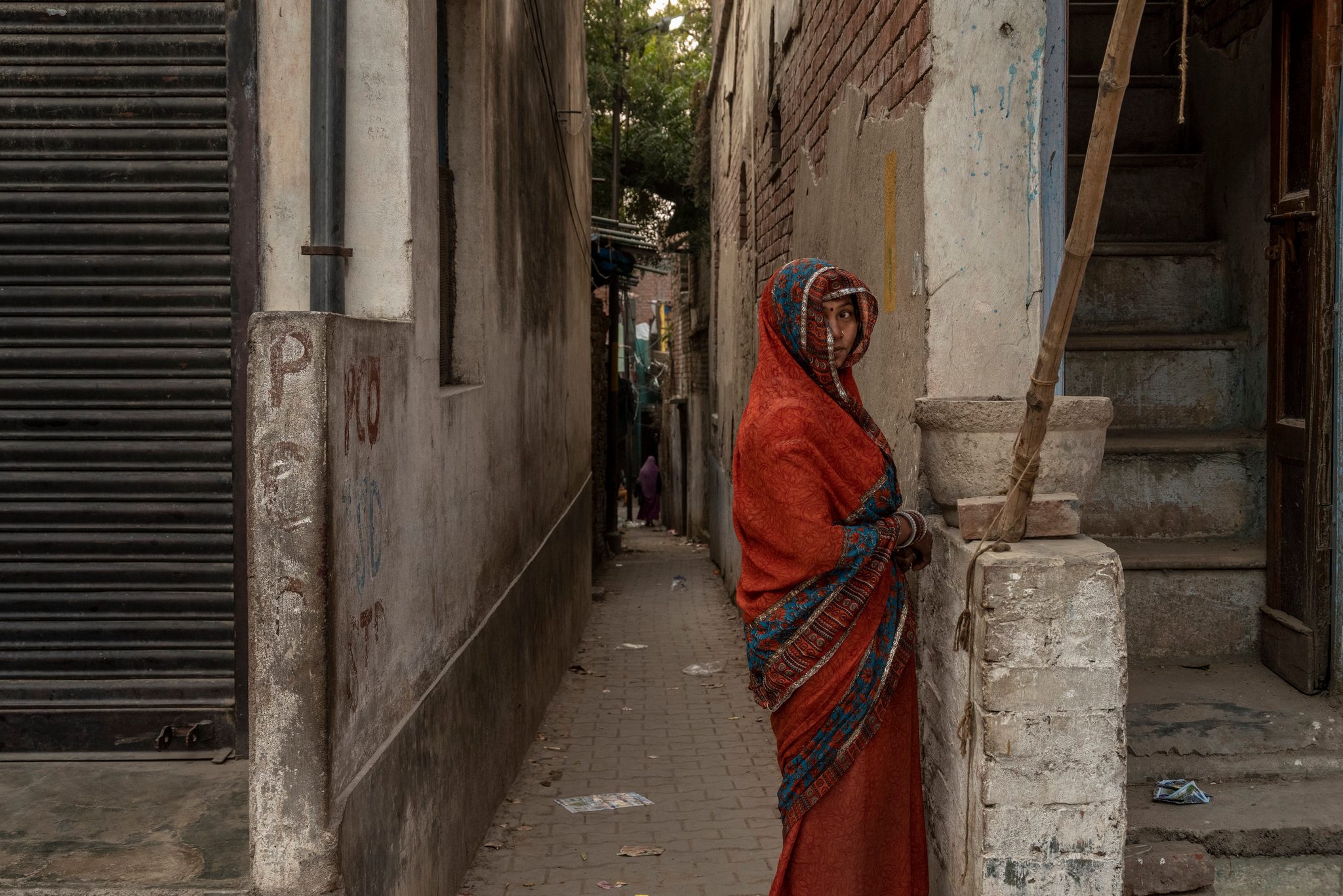 A woman in orange waits at the mouth of a narrow lane, one hand on the gatepost.
