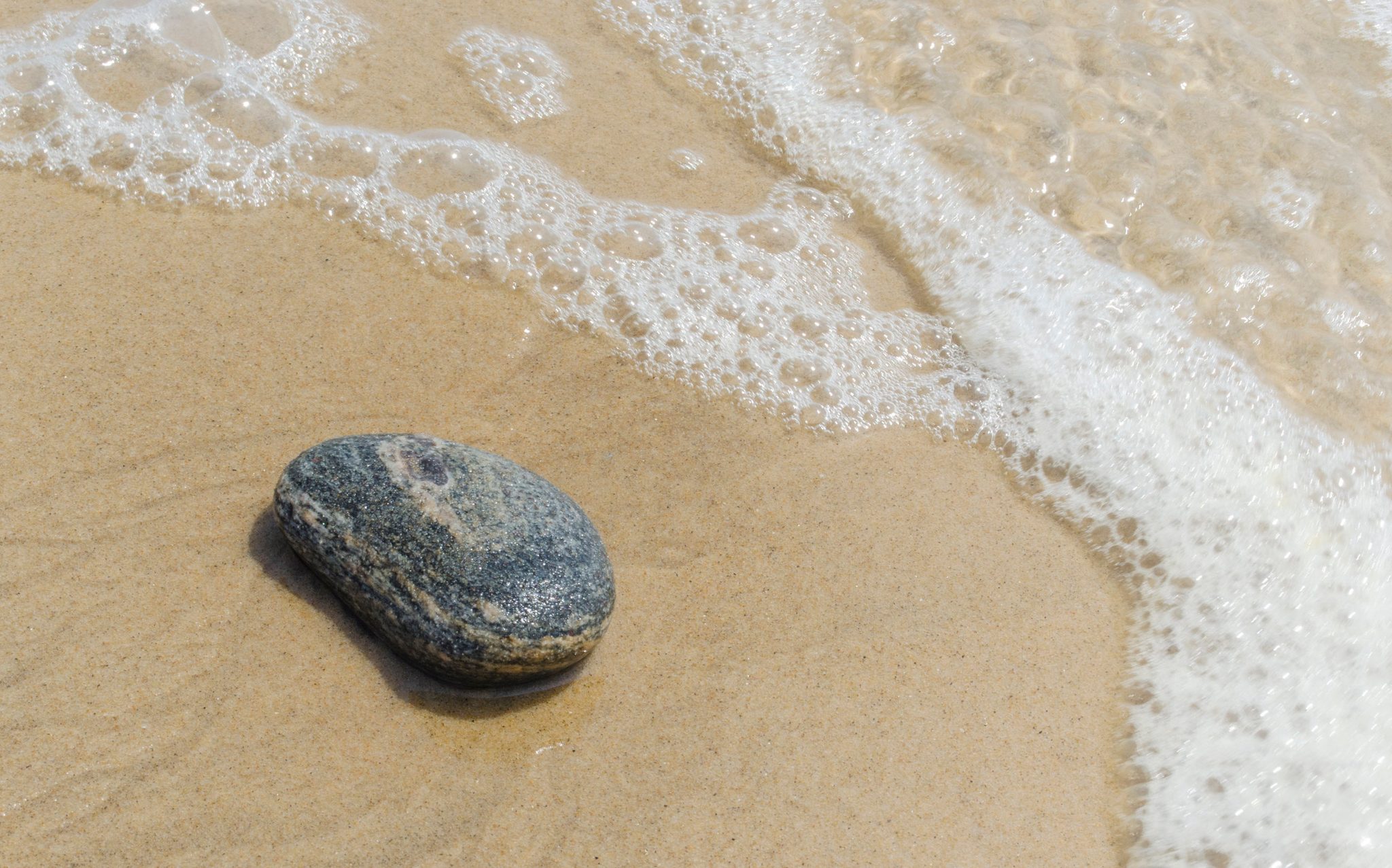 A single stone waits on the sand as the foam washes in.