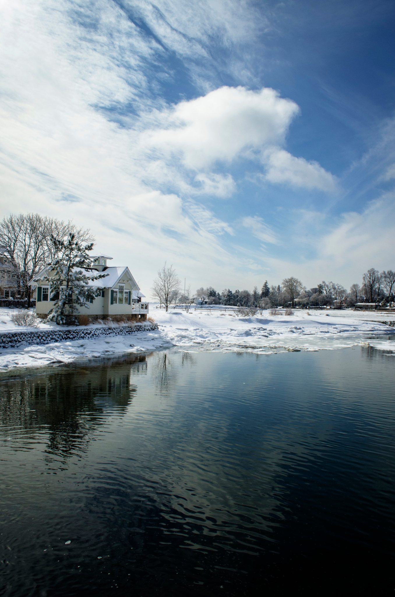 A white house sits by the dark water, snow on the banks and clouds overhead.