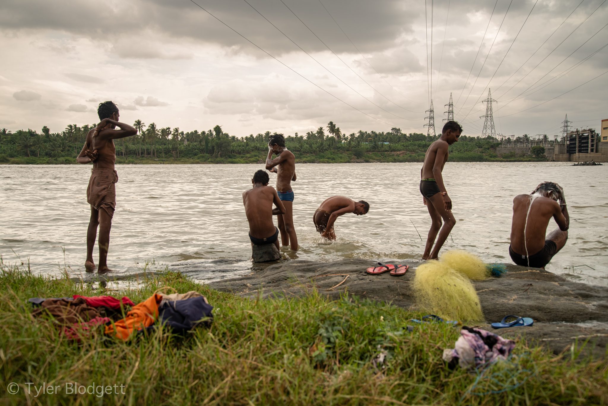 Men bathe at the river's edge beneath the power lines, a yellow net drying on the rocks.