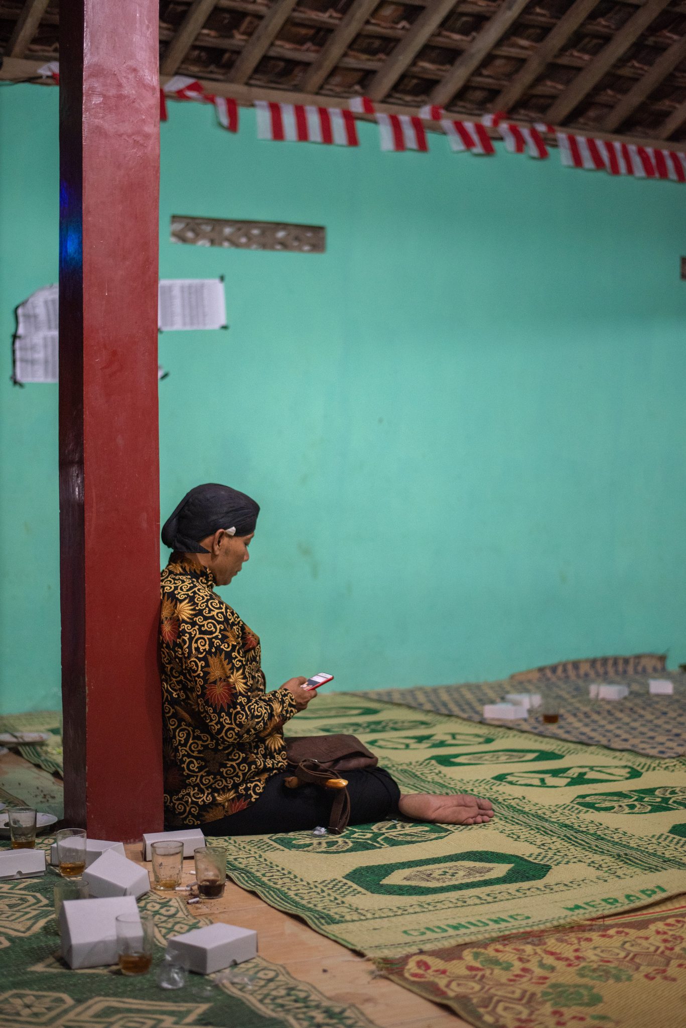 A man in batik leans against a red pillar with his phone, red and white bunting strung overhead.