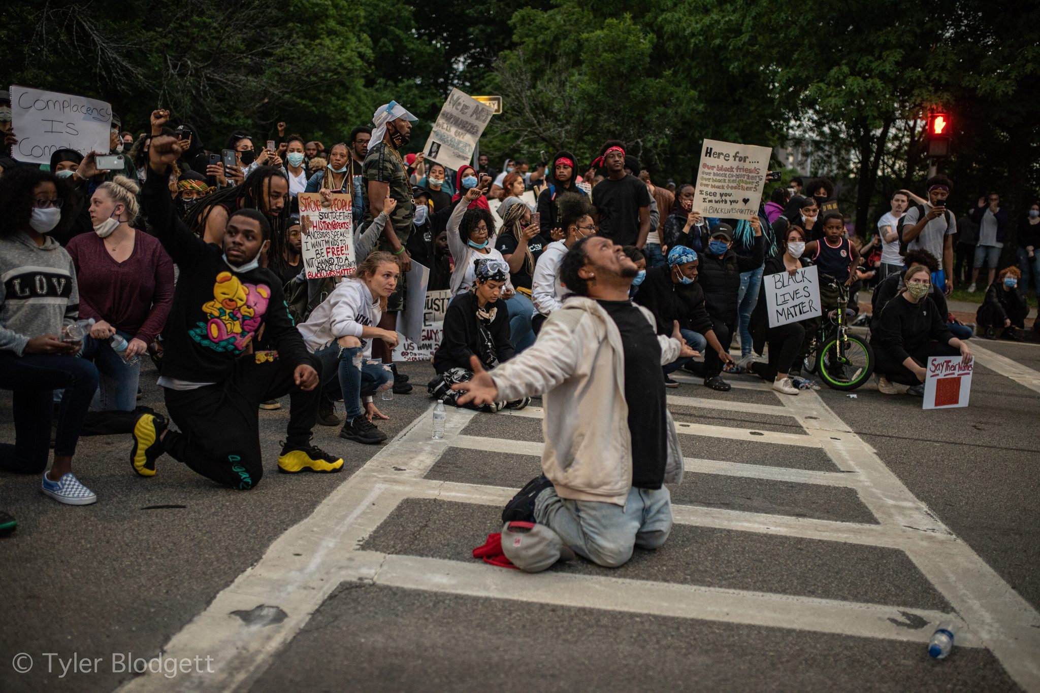 A man kneels in the crosswalk, arms open and face to the sky. Around him, the crowd kneels too.