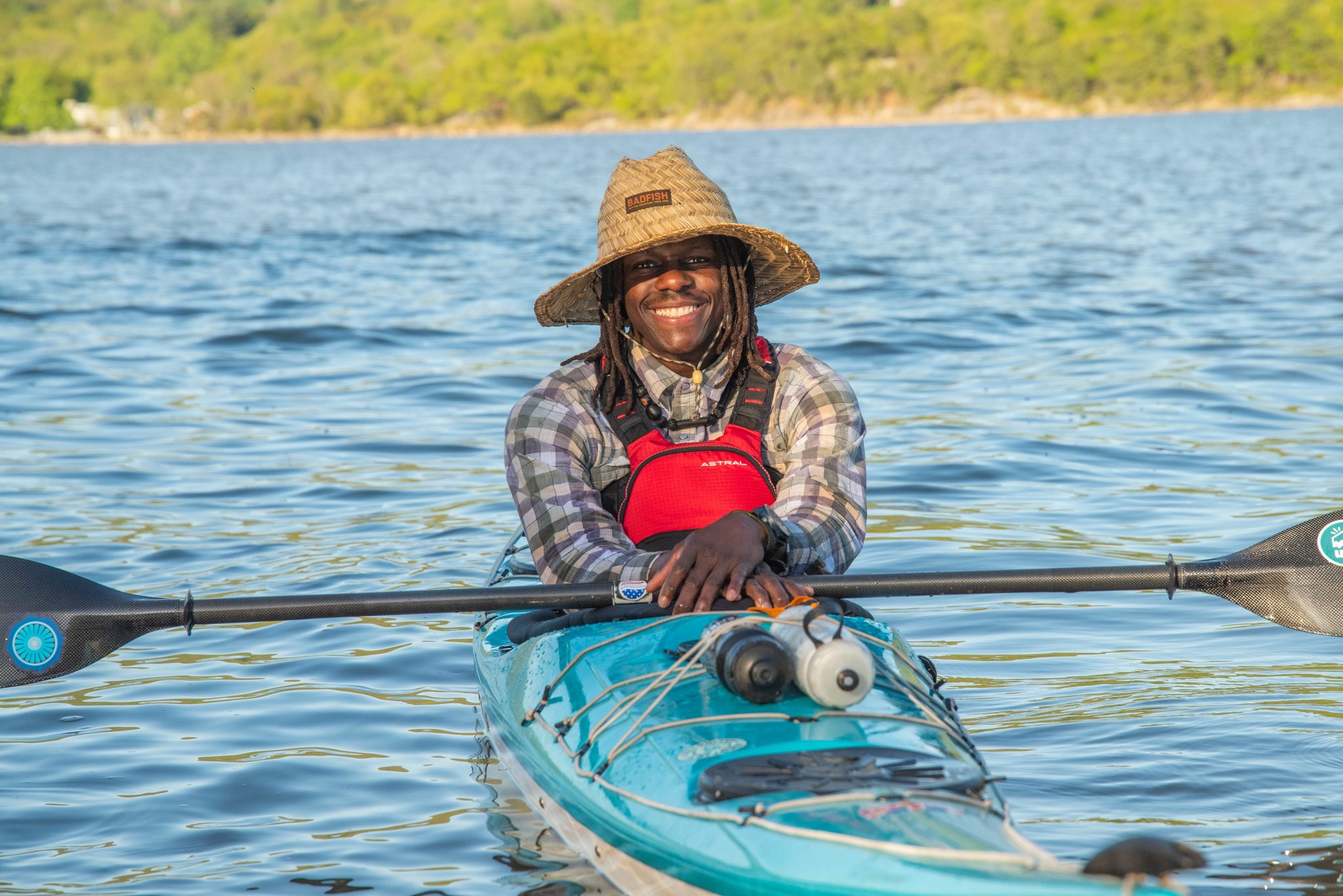 Straw hat, red life vest, both hands on the paddle shaft. Chev sits easy in the kayak on bright blue water.