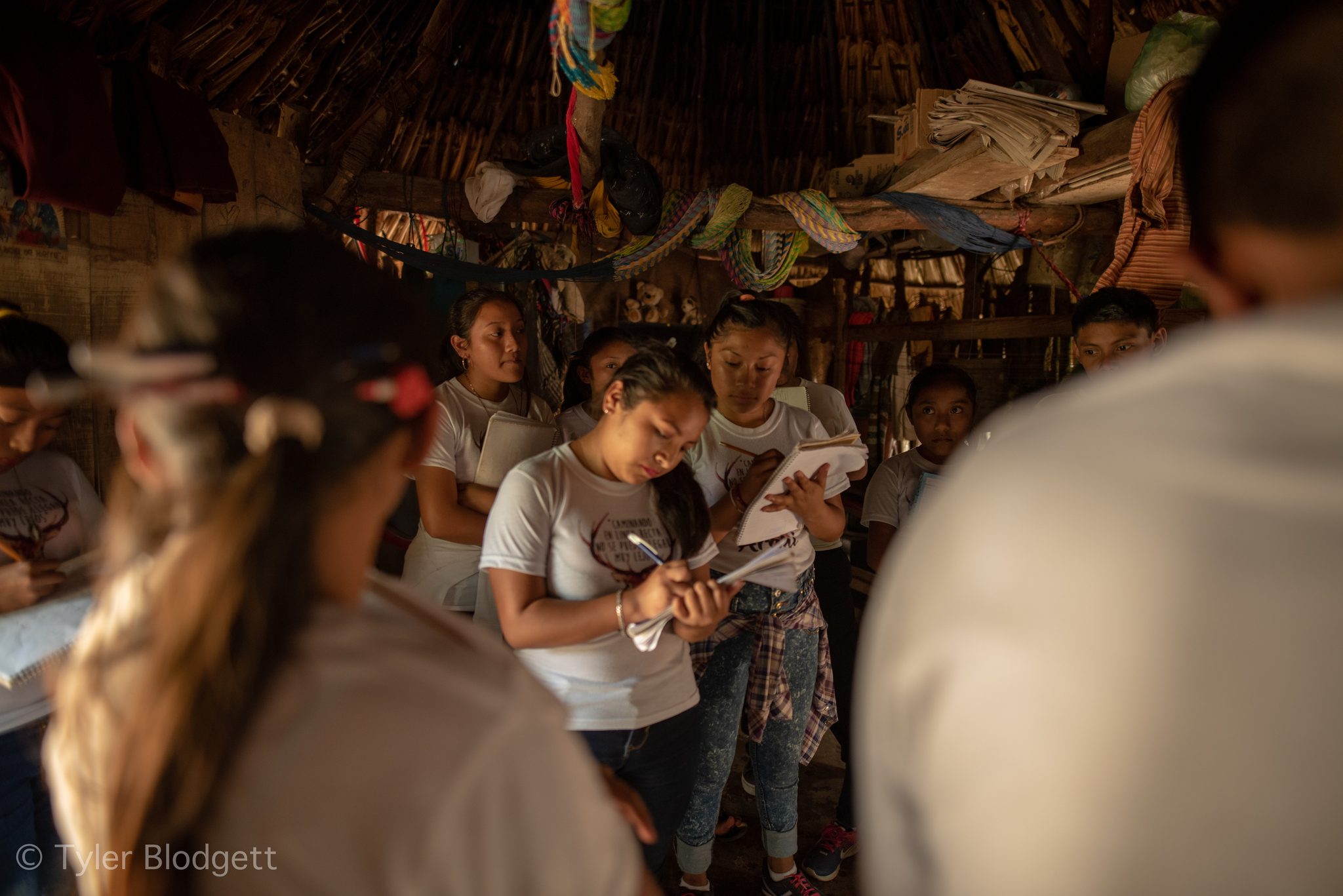 Students take notes beneath a thatched roof hung with bags and papers.