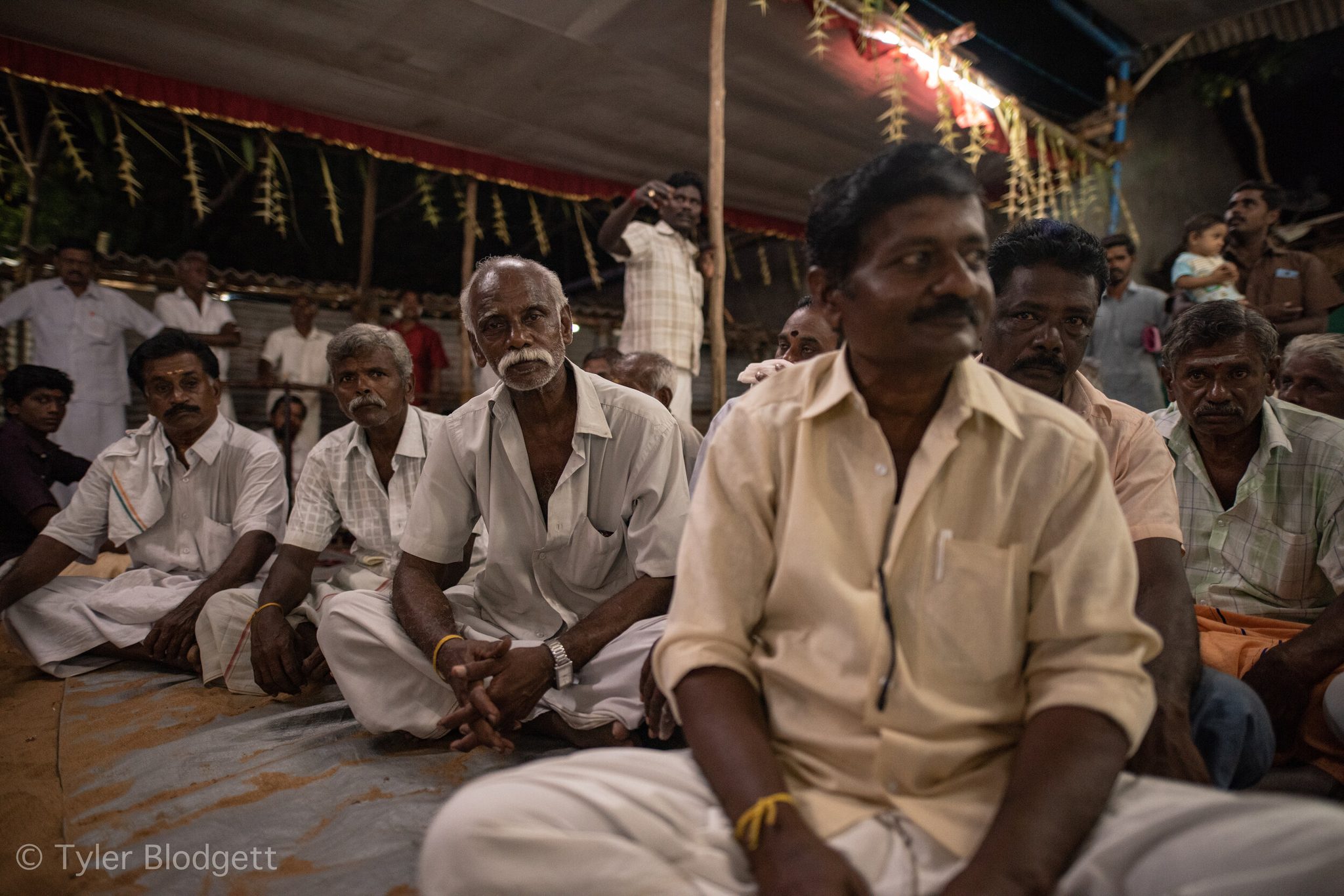 Men sit shoulder to shoulder on the ground beneath a festival canopy, every eye on something just out of frame.