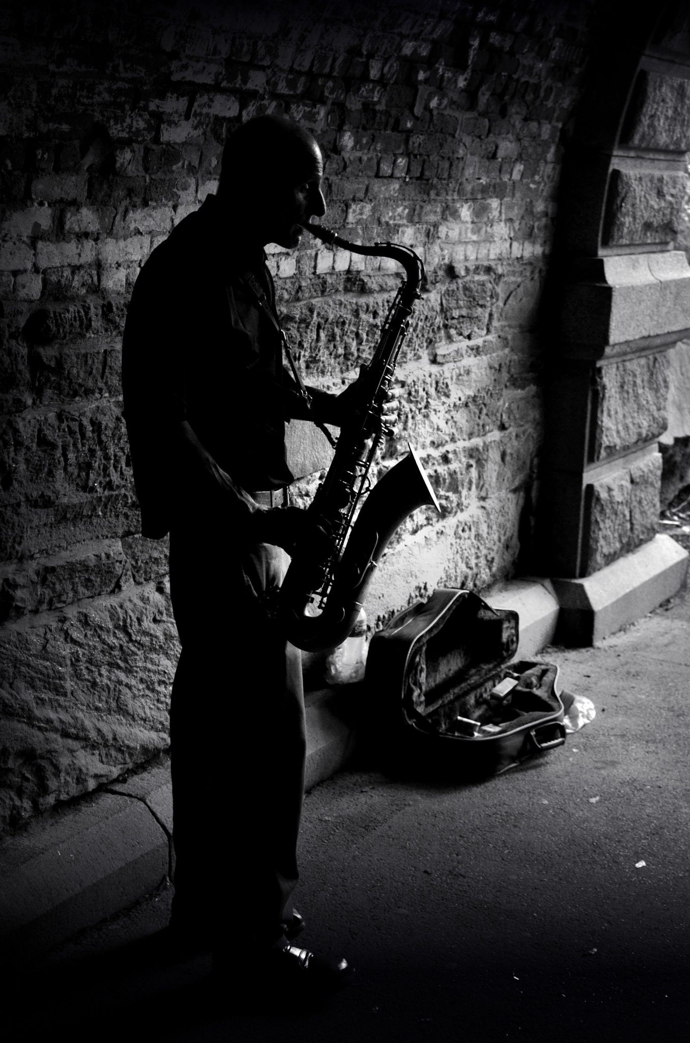 A saxophonist plays in the shadow of the stone arch, his case open at his feet.