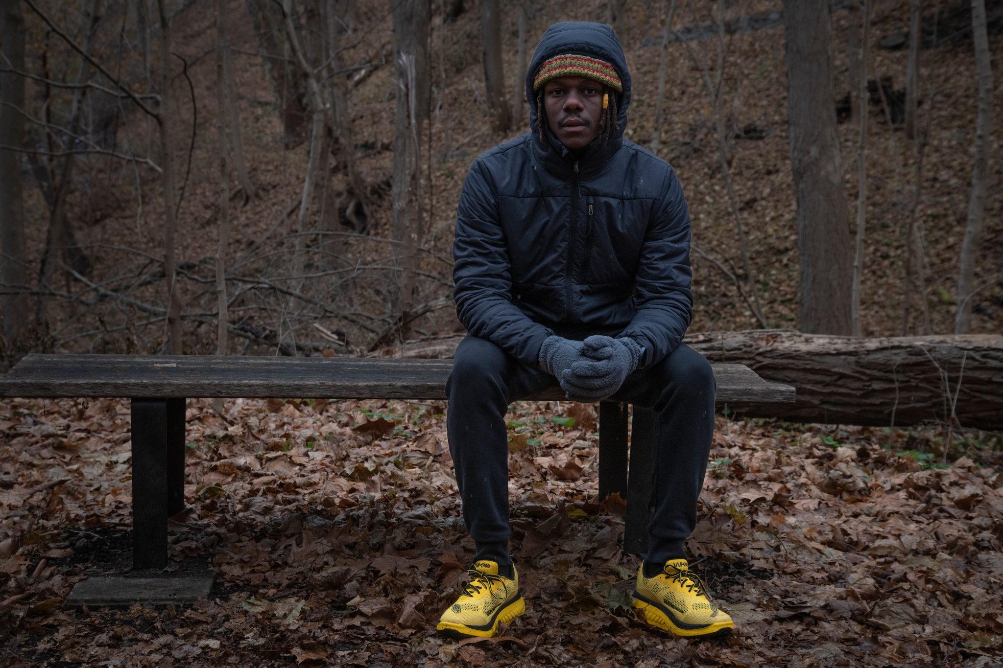 Chev rests on a log bench in the leaf litter, gloved hands folded, shoes bright against brown woods.