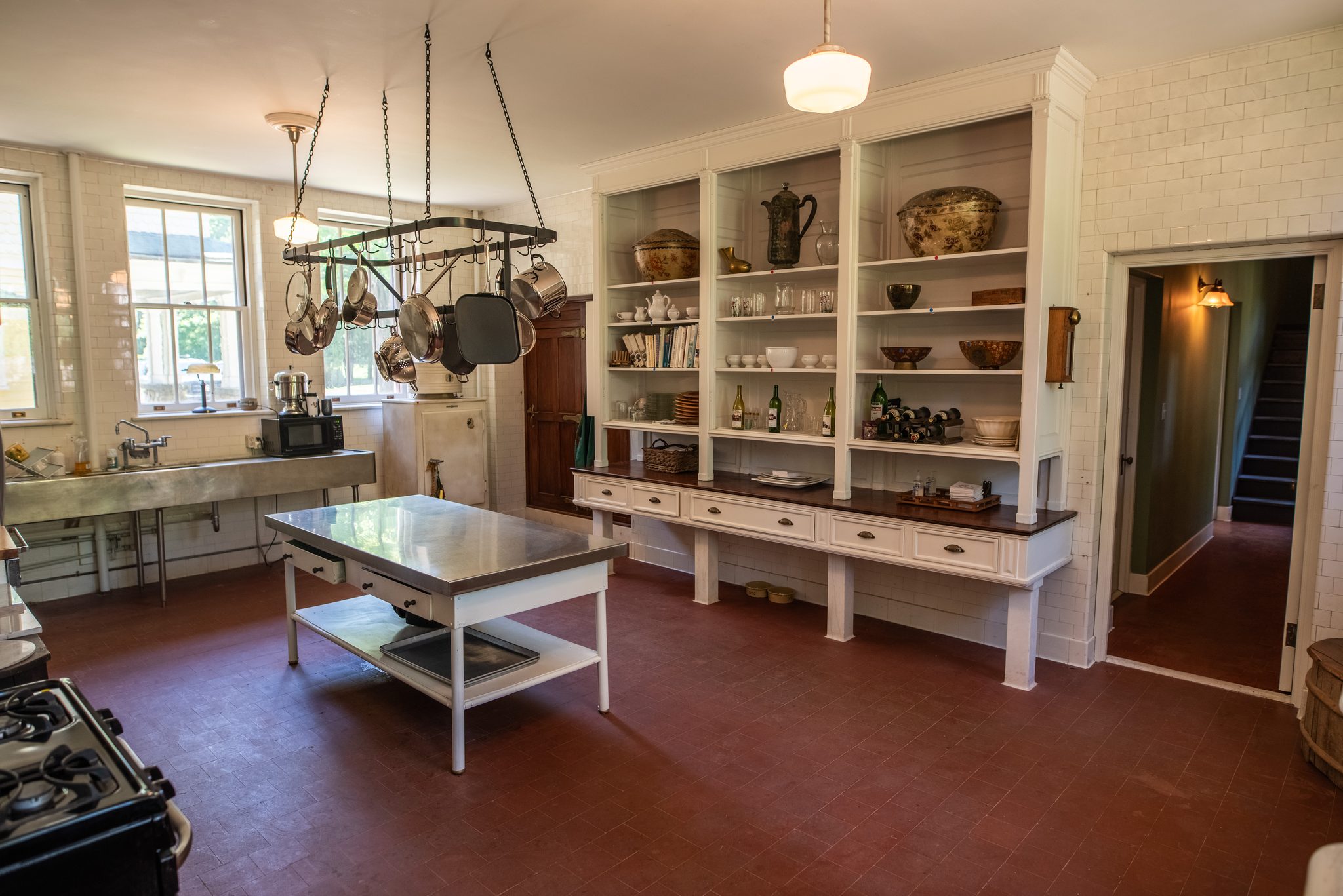 The kitchen keeps its white subway tile, open shelving, a steel-topped work table and a rack of hanging copper pans.
