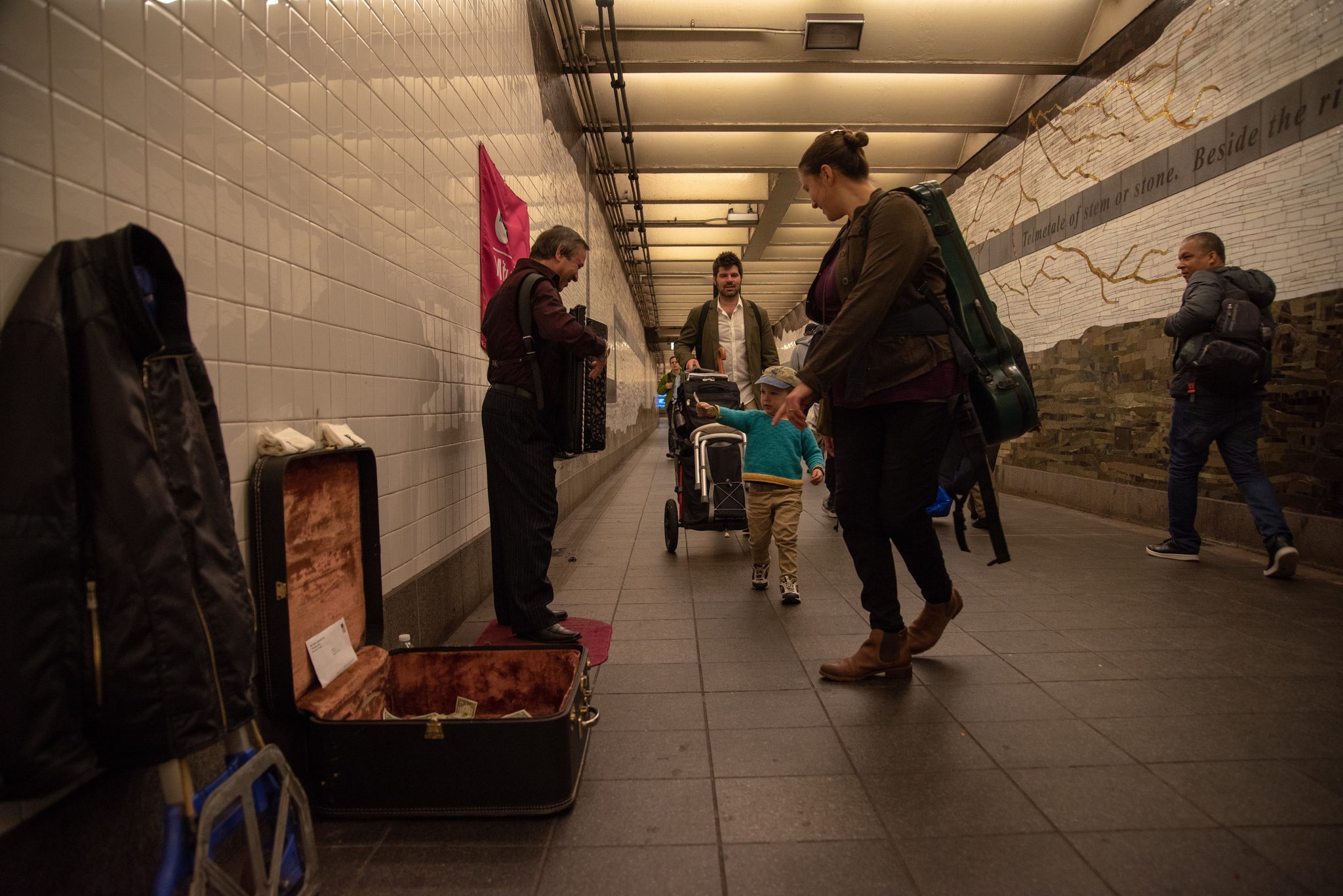 In a tiled corridor, the family passes another busker playing accordion over his open case.