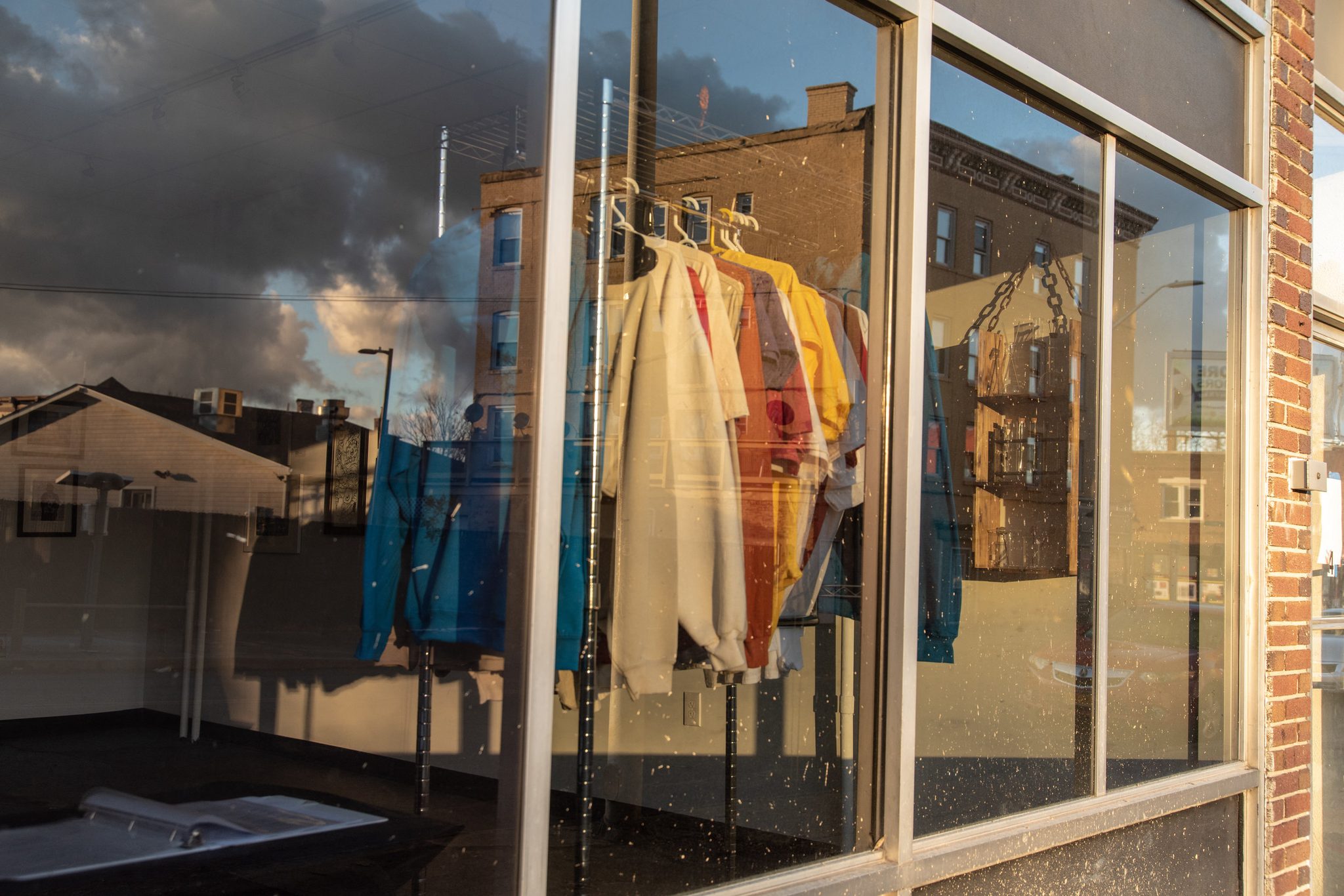 Coats hang in a shop window as the clouds and brick buildings reflect across the glass.