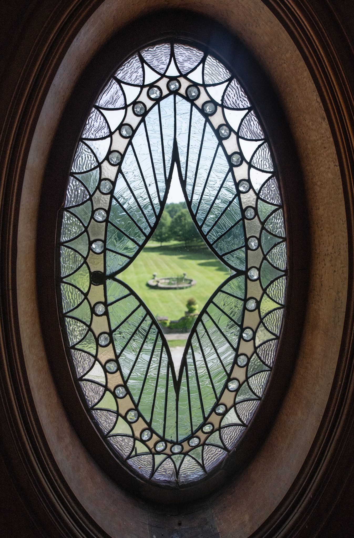 An oval leaded window frames the lawn and its round stone fountain, seen from high up inside the house.