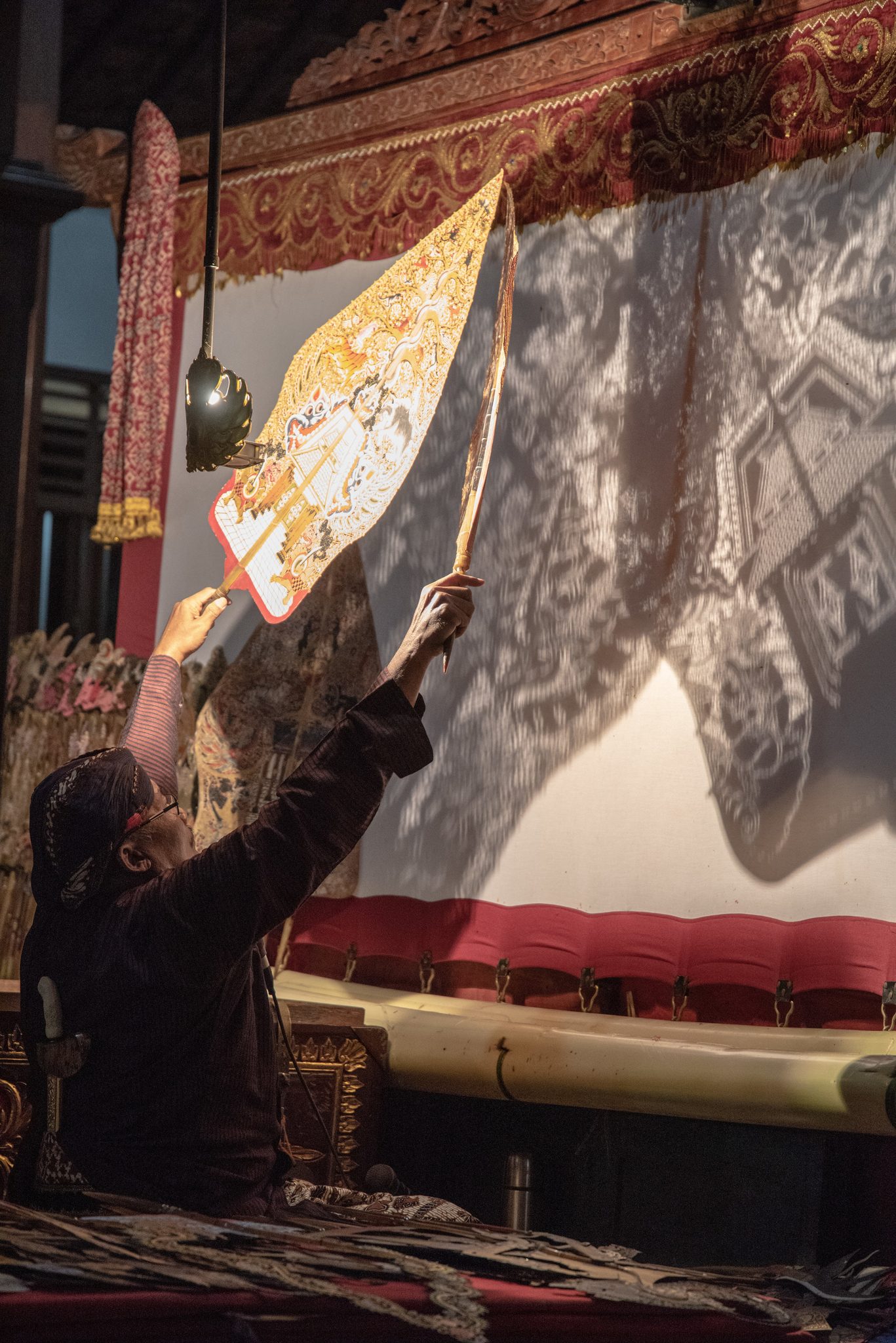 Ki Kasidi raises the gunungan to the screen, and its shadow spreads across the cloth.