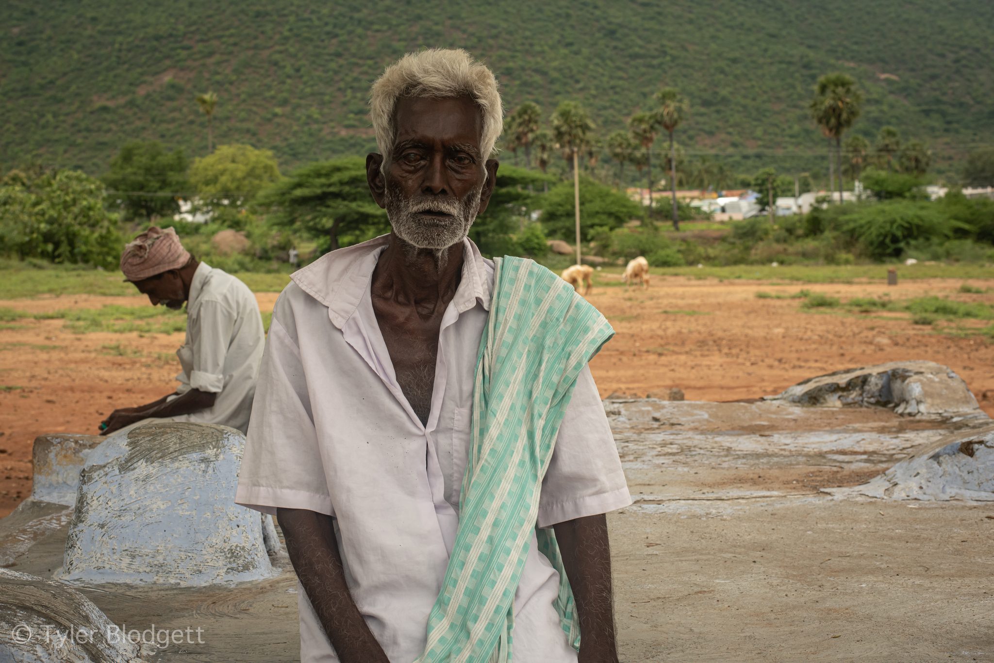 An elder in white, a green towel over his shoulder, stands at the edge of the clearing.