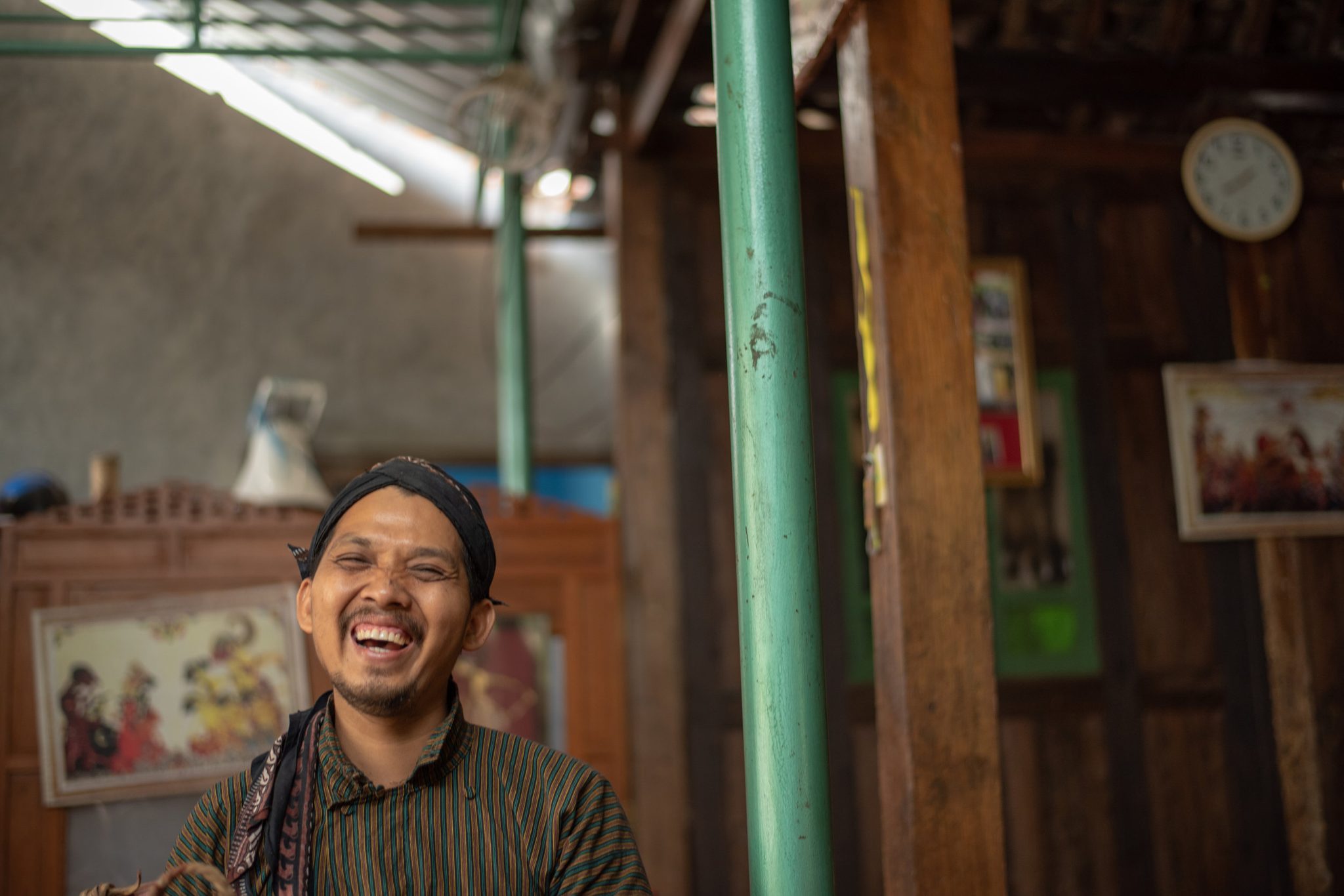 Dhidot throws his head back and laughs, the wall behind him hung with wayang paintings.