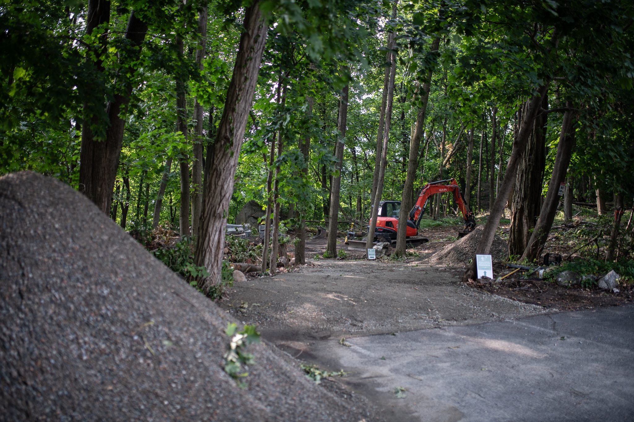 The old path runs past a pile of gravel toward the excavator waiting in the trees.