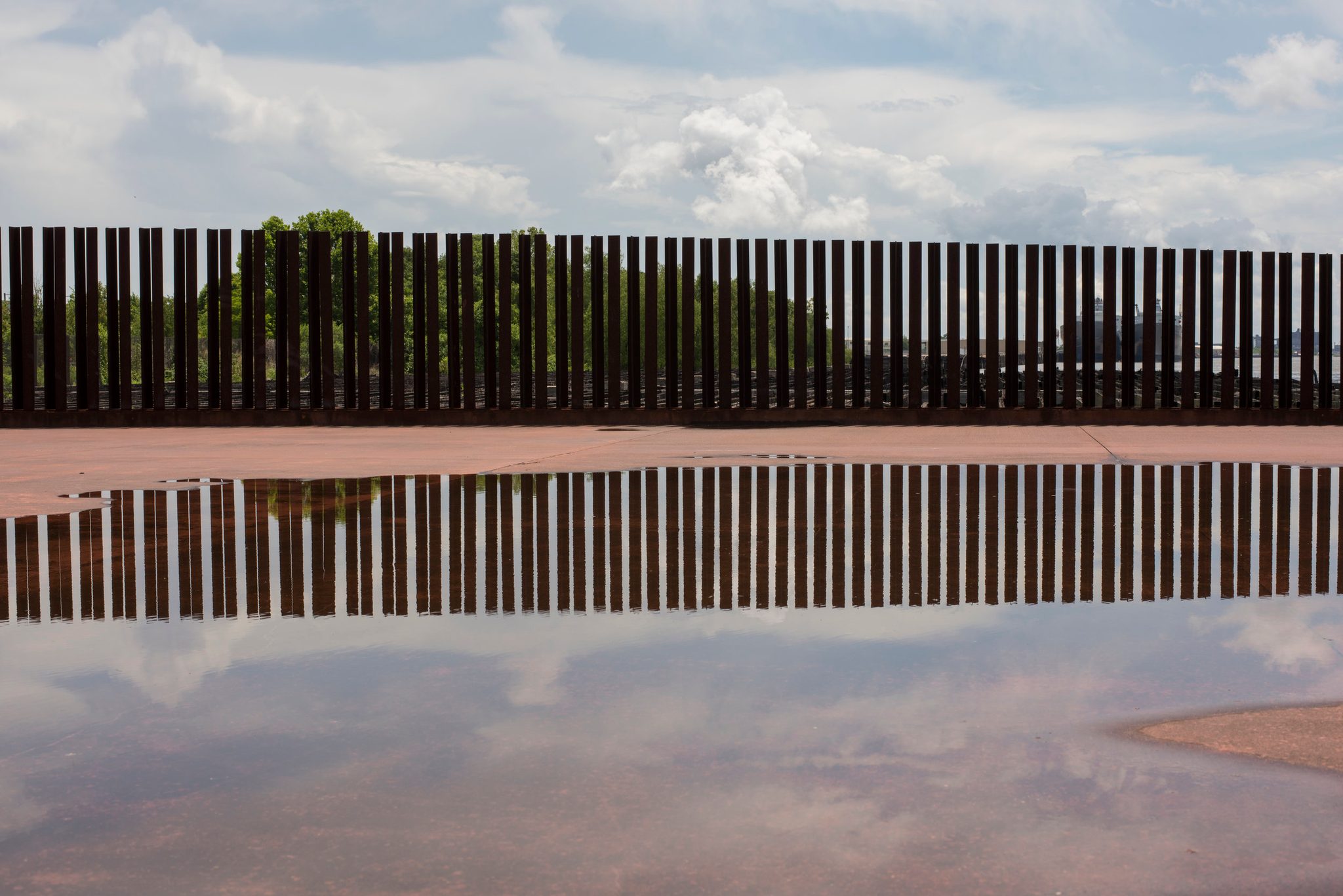 A rust-red fence and the clouds above it, doubled in a rain puddle.