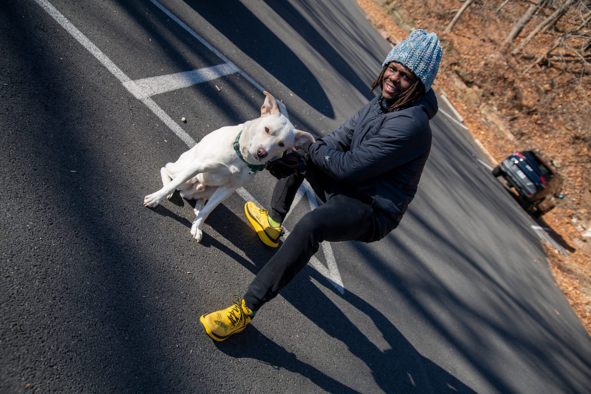 A white dog leaps up for play on the road and Chev leans back laughing, shoes planted.