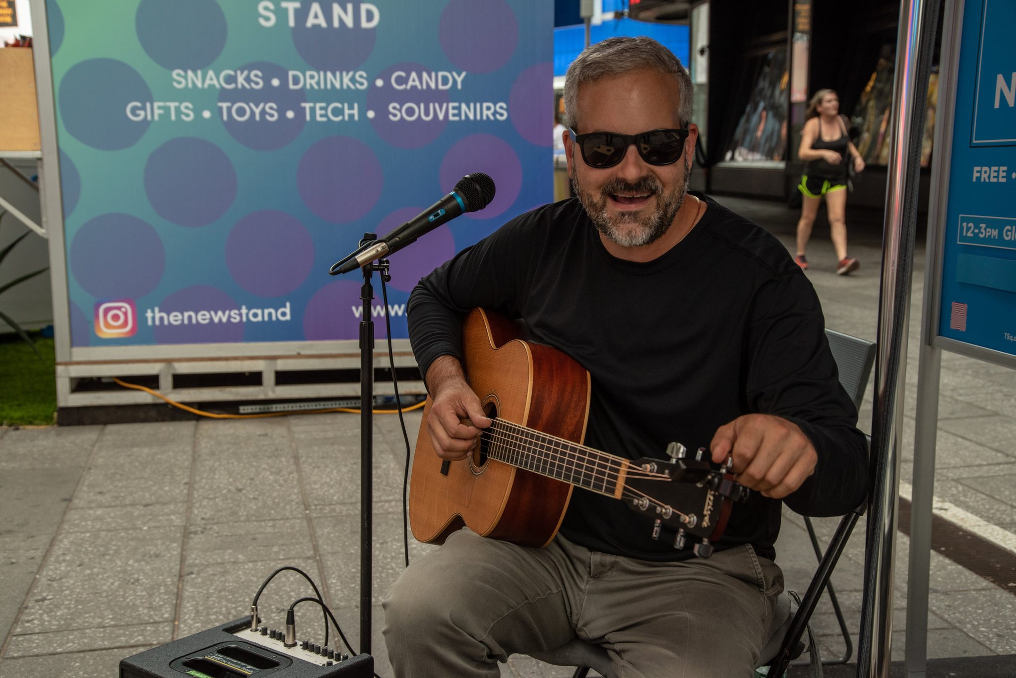 Glenn grins behind his sunglasses, guitar in his lap, set up beside a Times Square newsstand.
