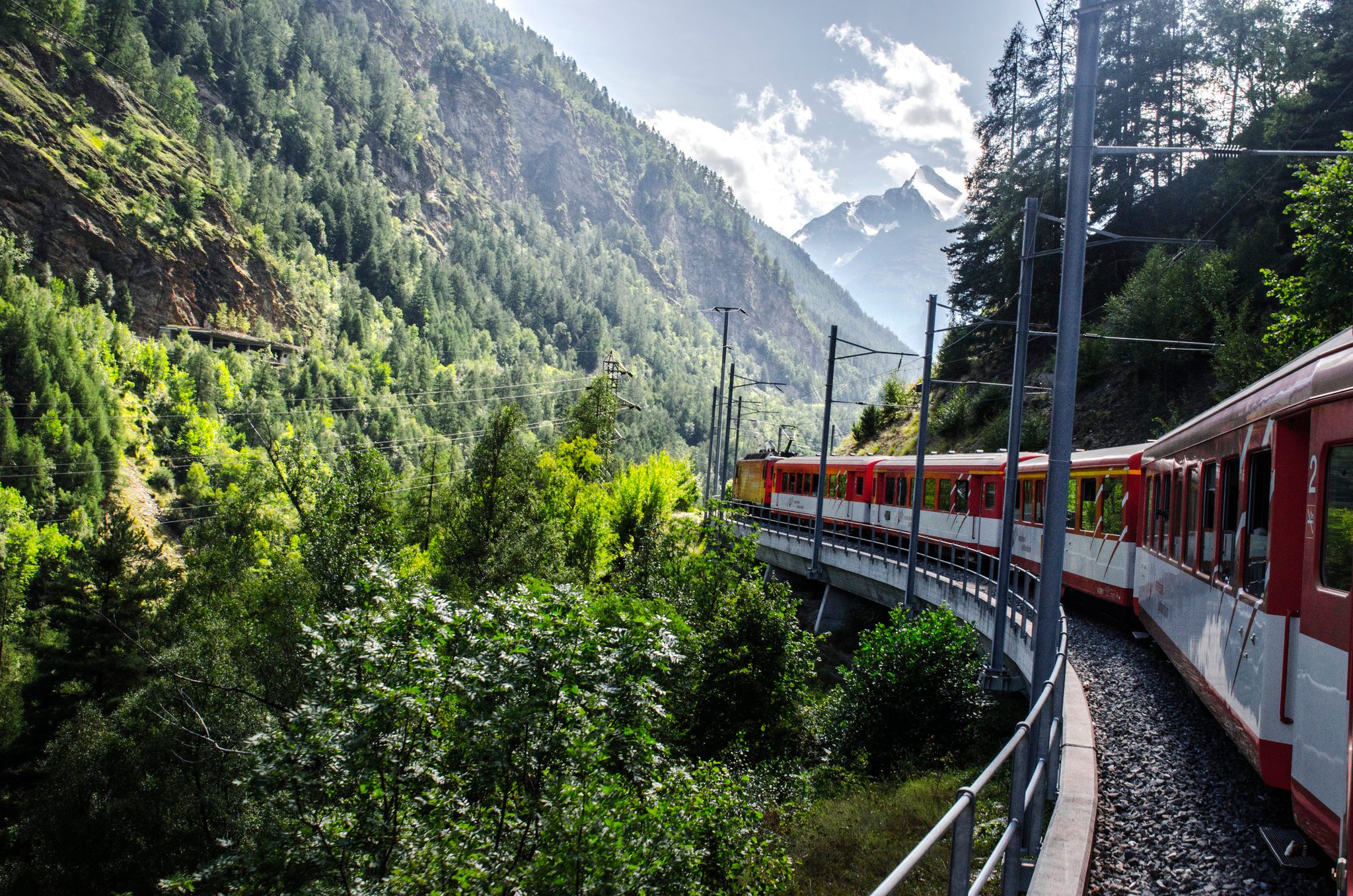 The red train curves along the mountainside toward the snowy peaks.