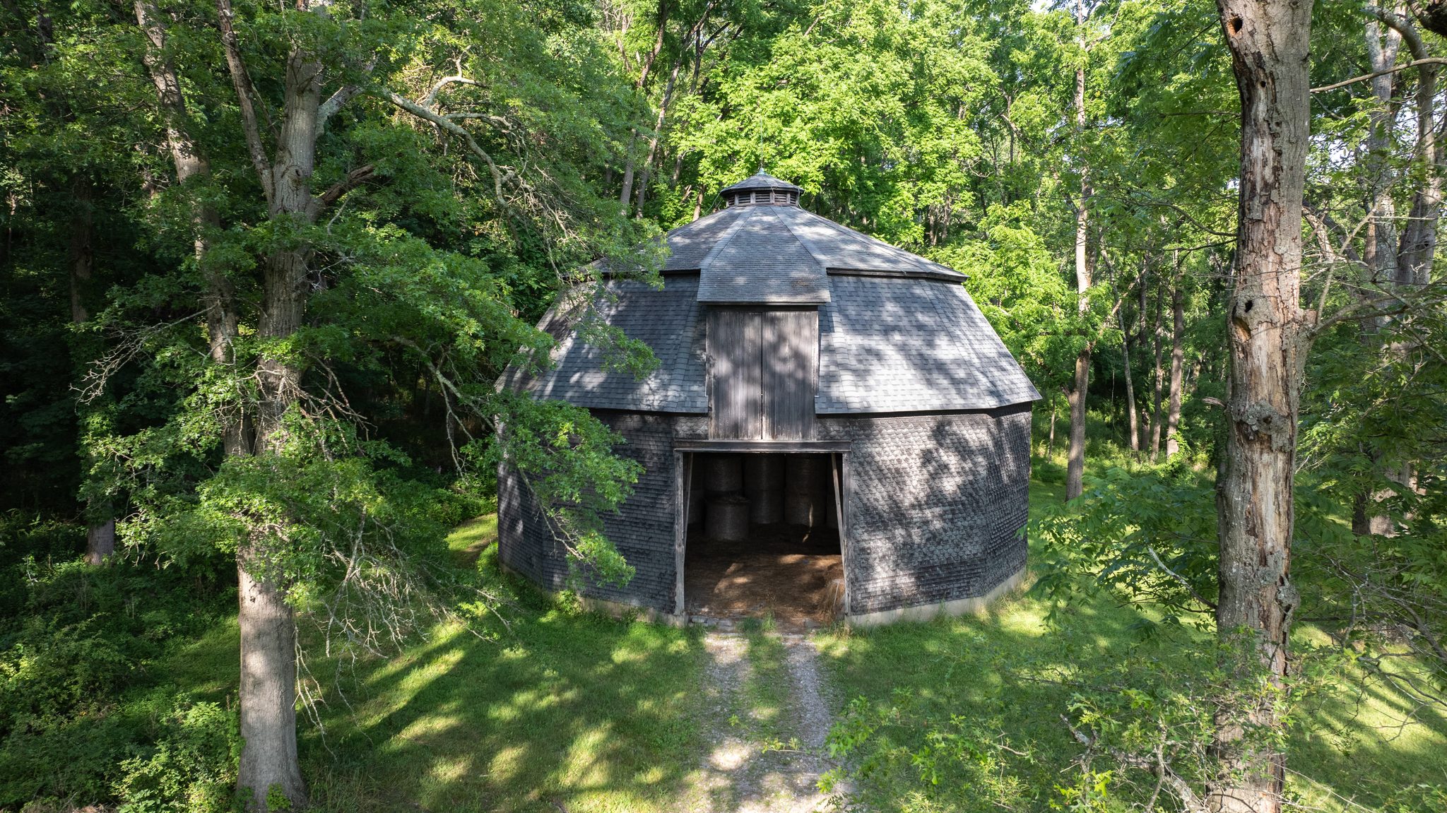 A shingled polygonal barn in the woods, its wide door open onto round bales stacked inside.
