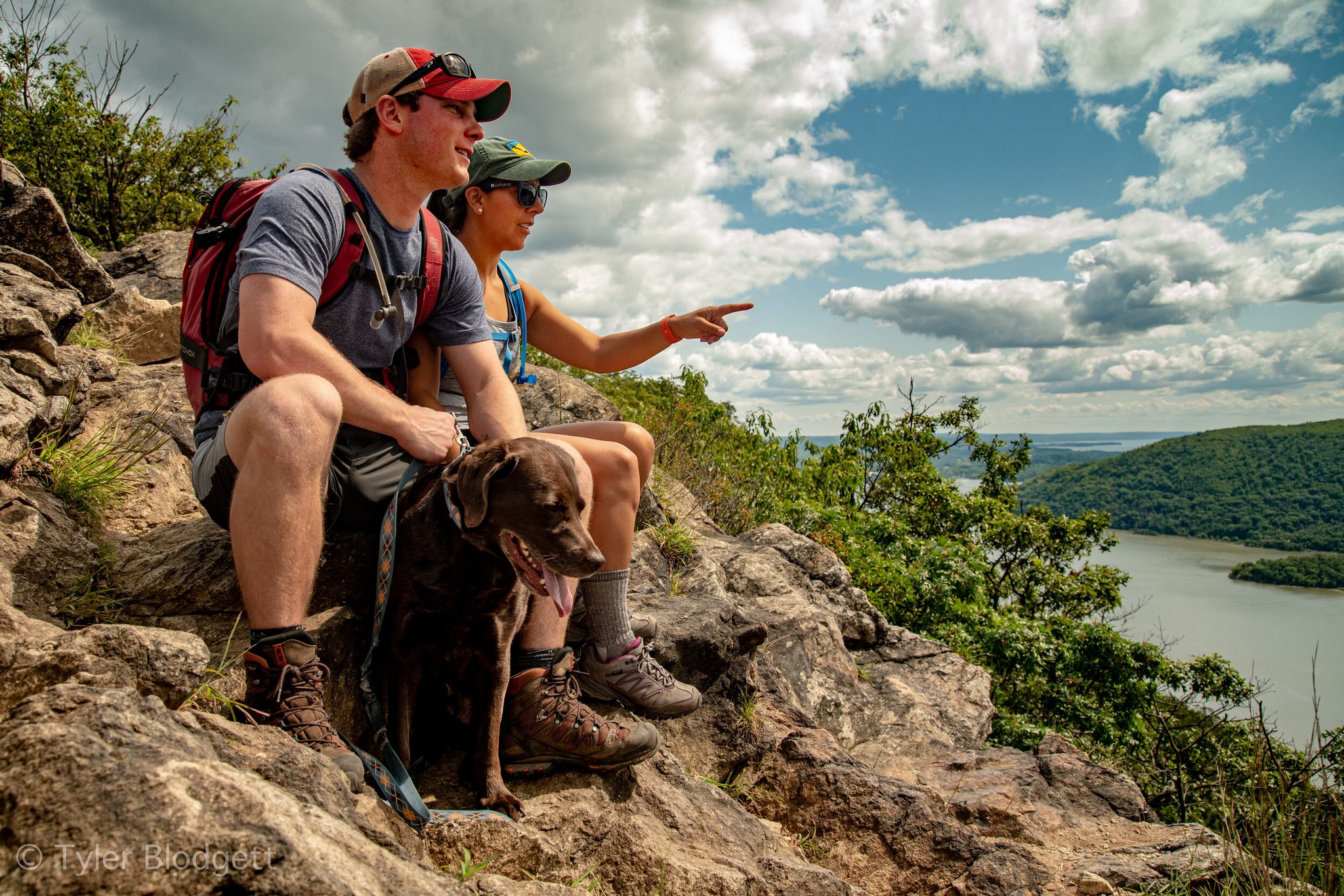 A pair of hikers and their chocolate lab stop on the rocks, one of them pointing across the river.