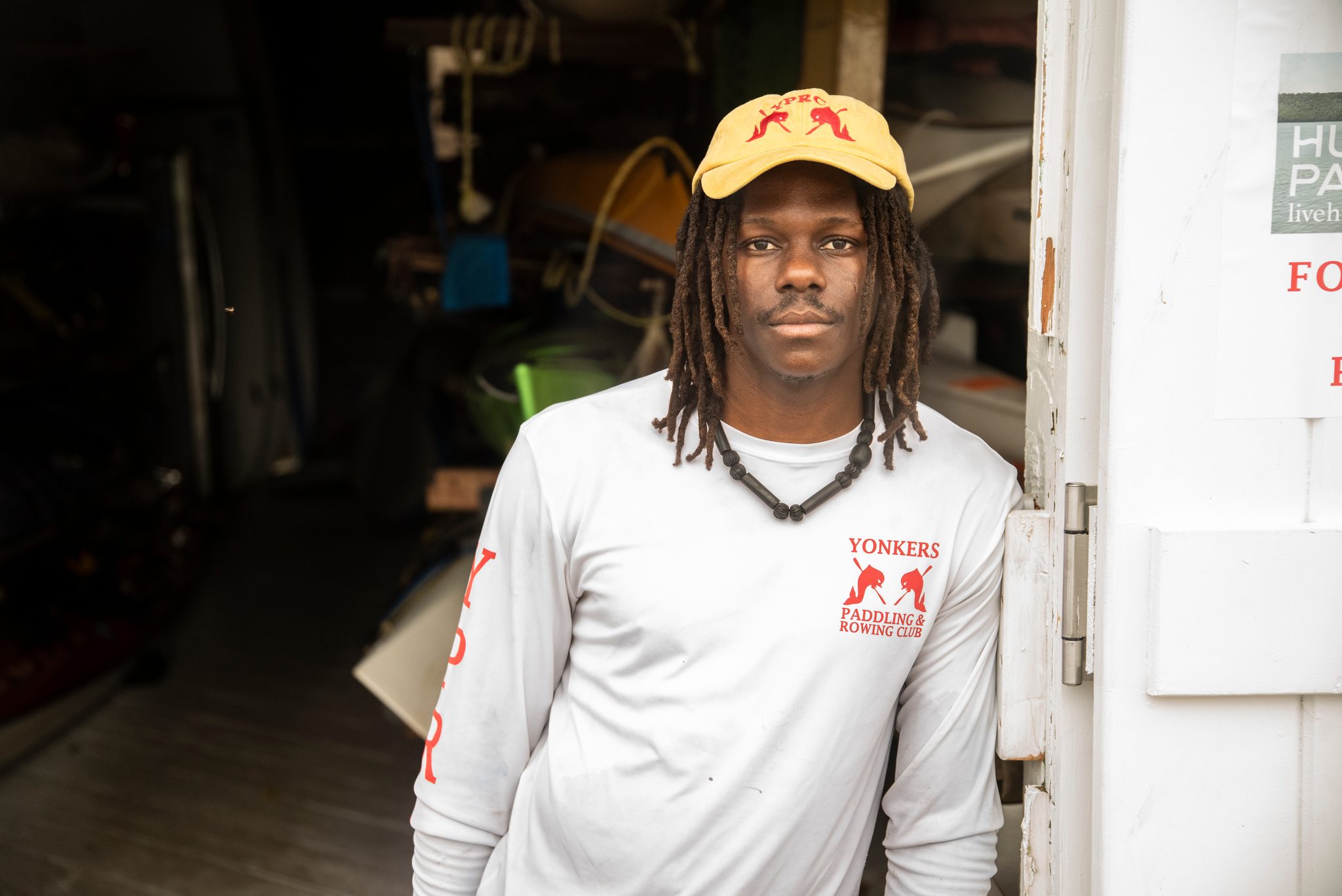 Chev in the boathouse doorway, yellow cap and paddling club shirt, a quiet moment between legs.