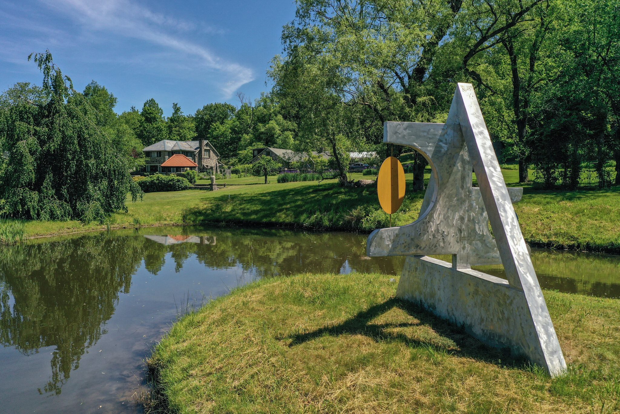 A steel sculpture stands on a point of grass at the edge of the pond.