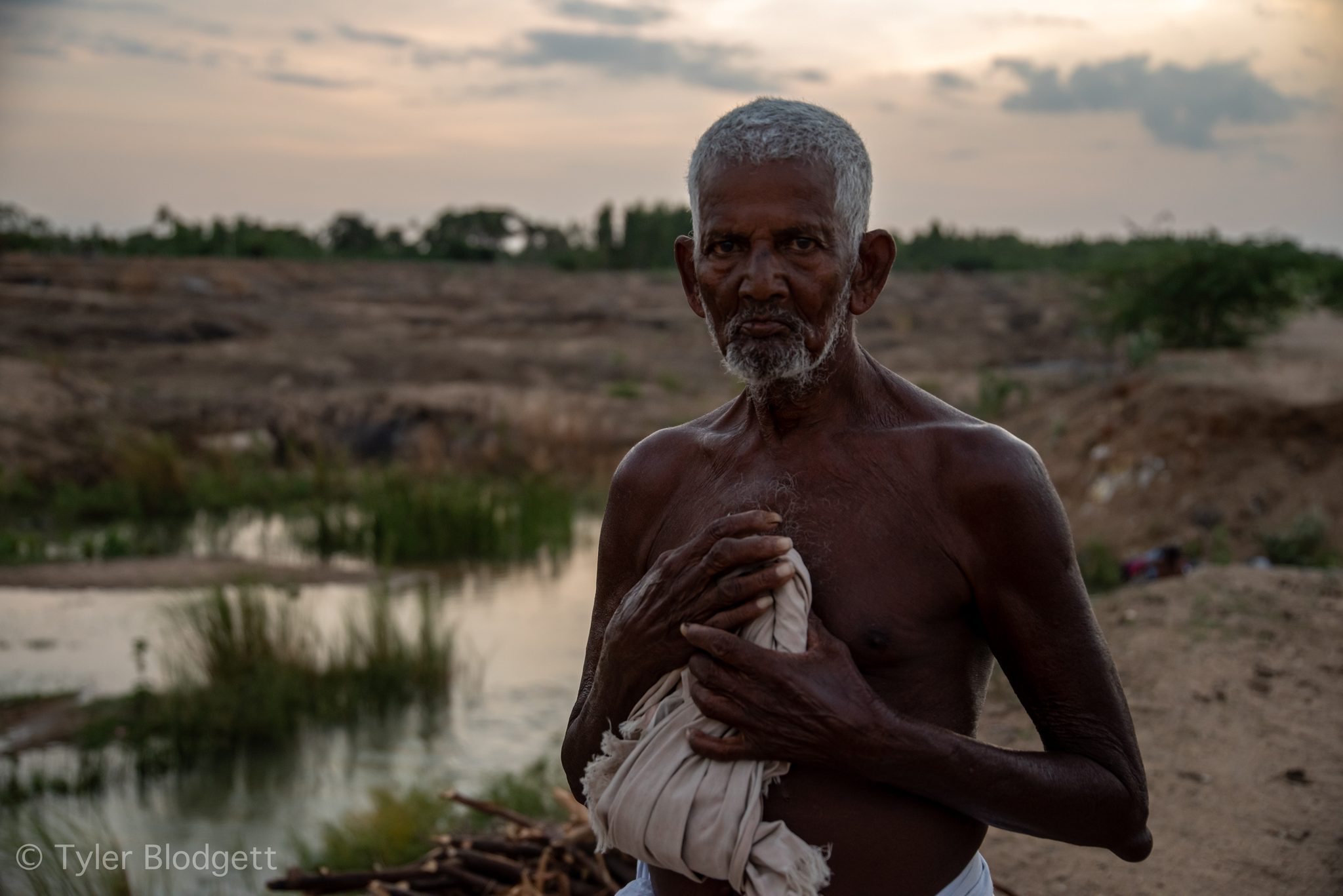 An elder holds a cloth to his chest as the sun goes down behind him.