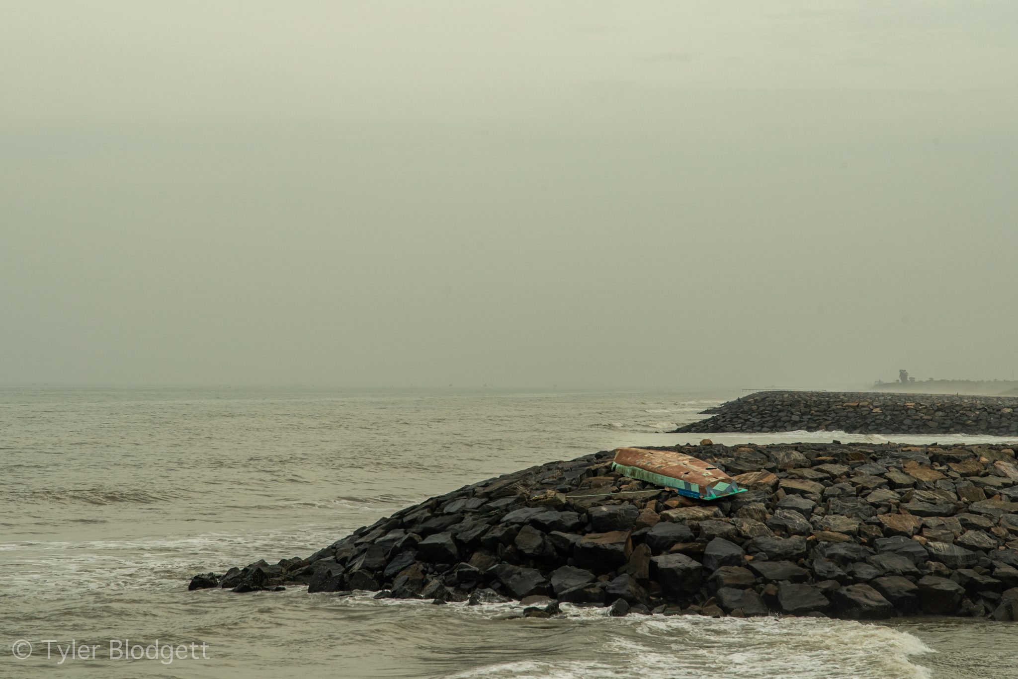 Waves wash against a jetty of dark boulders under a white sky.