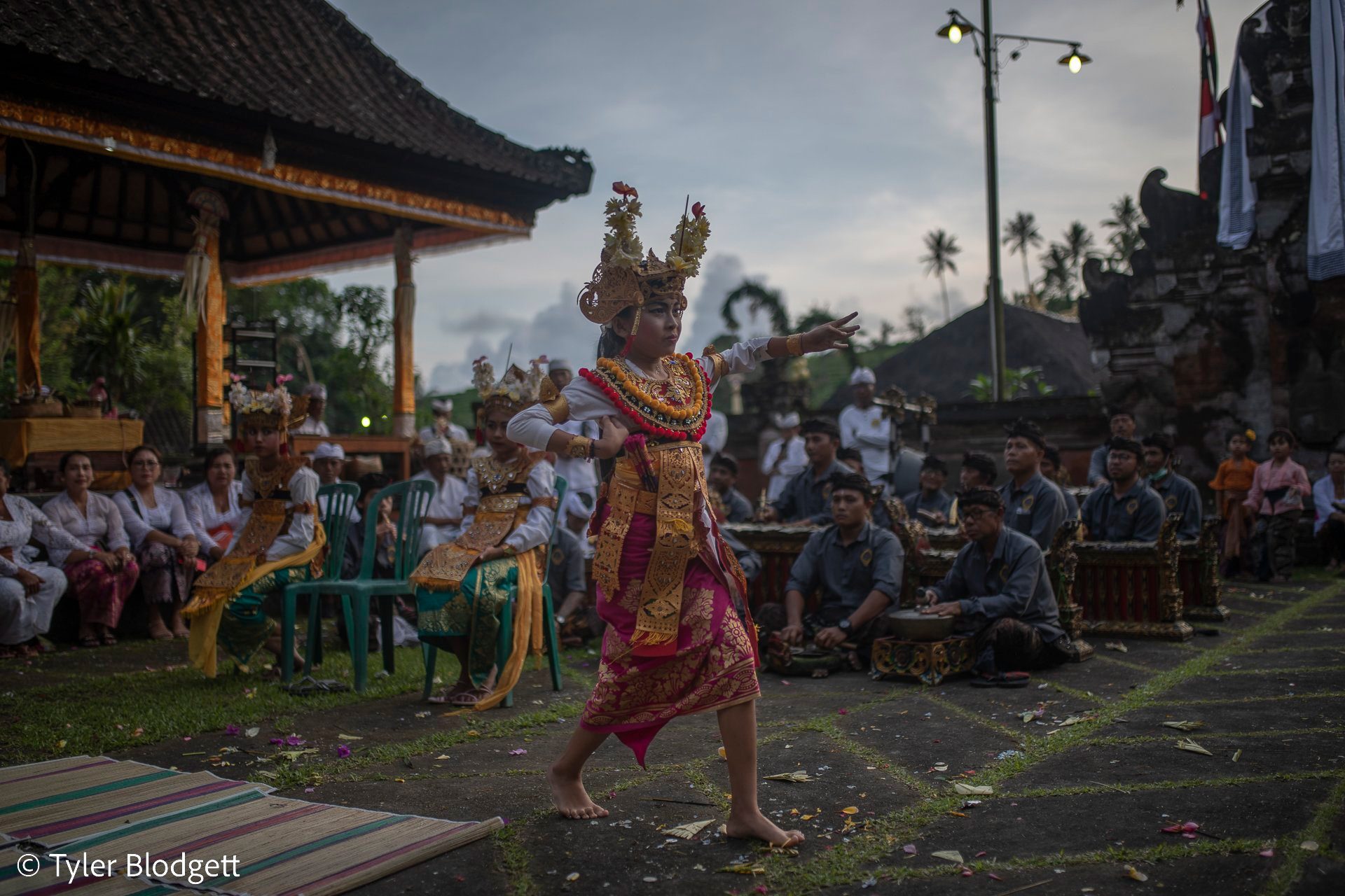 A dancer in gold and a tall crown steps across the grass as the crowd watches at dusk.